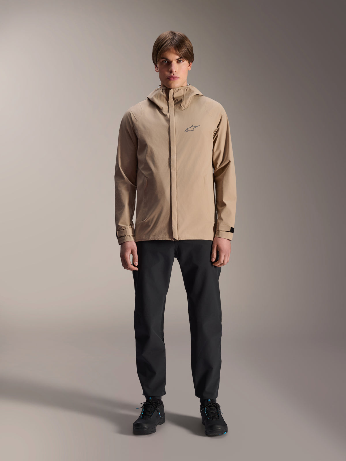 Veste de pluie Alpinestars A-Dura, veste de cyclisme brun clair, portée avec un pantalon noir et des chaussures noires, dotée d'un petit logo sur la poitrine et d'une capuche, équipement de VTT professionnel pour rouler par tous les temps.