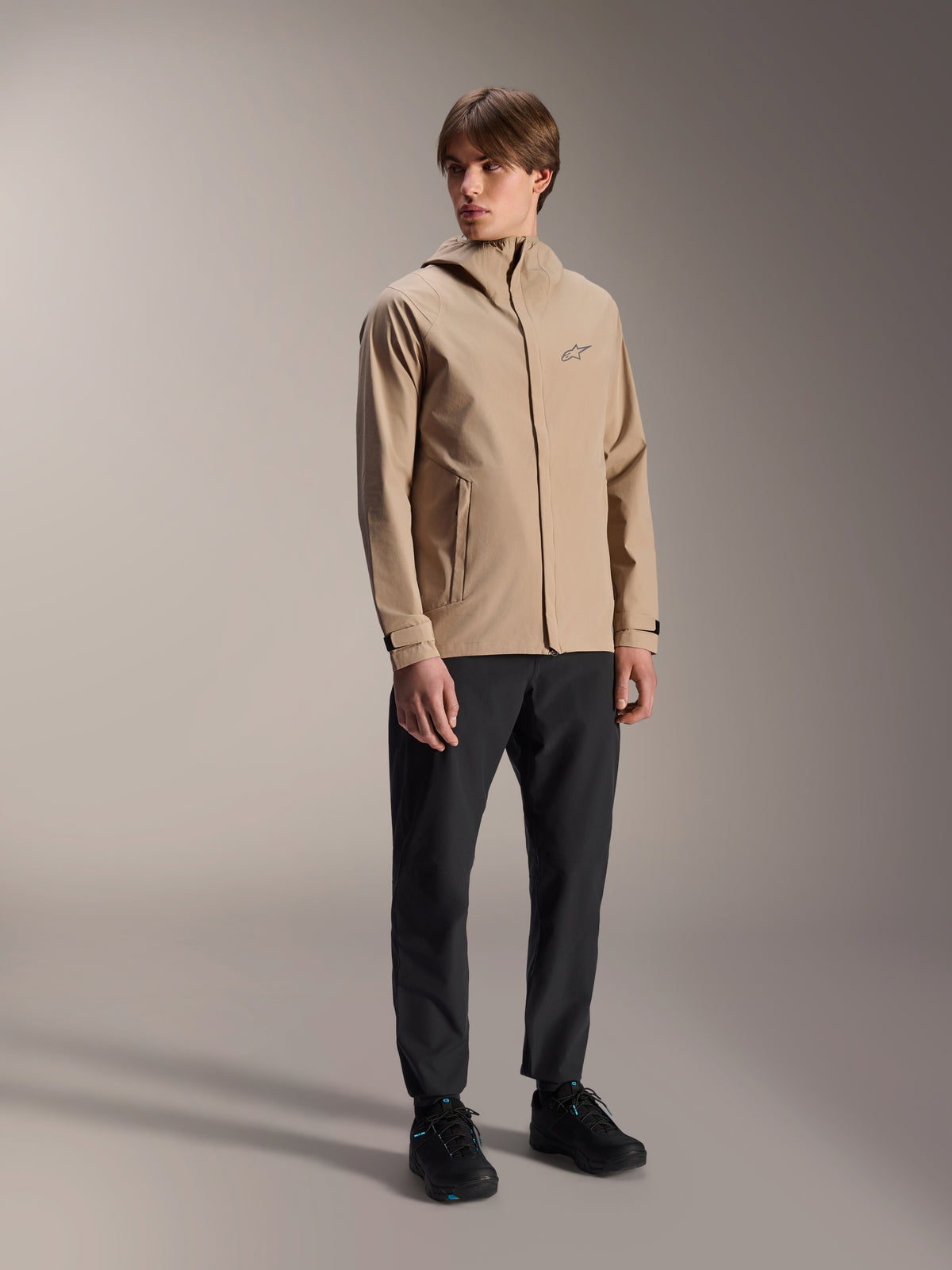 Modèle masculin portant la veste de pluie Alpinestars A-Dura en brun clair, avec un pantalon et des chaussures noirs, veste de pluie VTT au design minimaliste avec capuche et petit logo sur la poitrine.