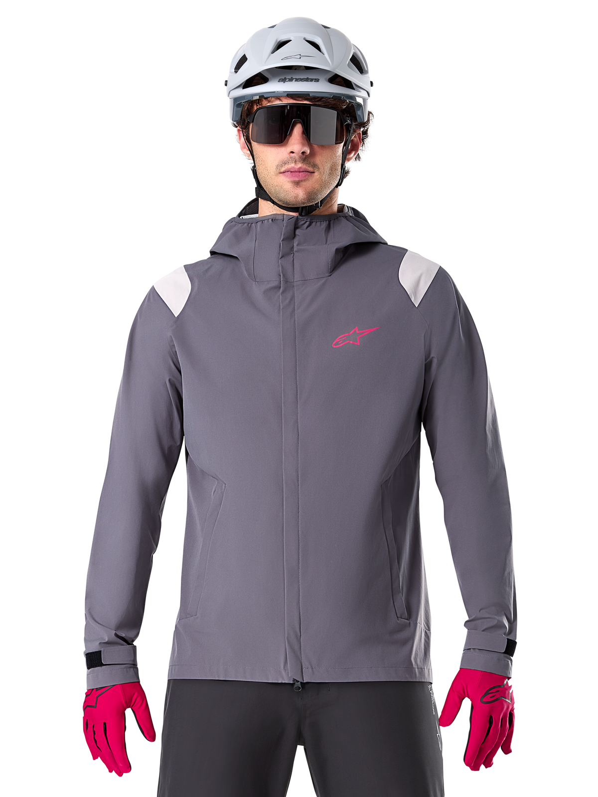 Alpinestars A-Dura Regenjas, mountainbike-regenjas, donker titaniumgrijs, ontwerp met capuchon, roze Alpinestars-logo en lichtgrijze schouderpanelen, gedragen met witte MTB-helm en felroze handschoenen, waterdichte uitrusting voor off-road fietsen