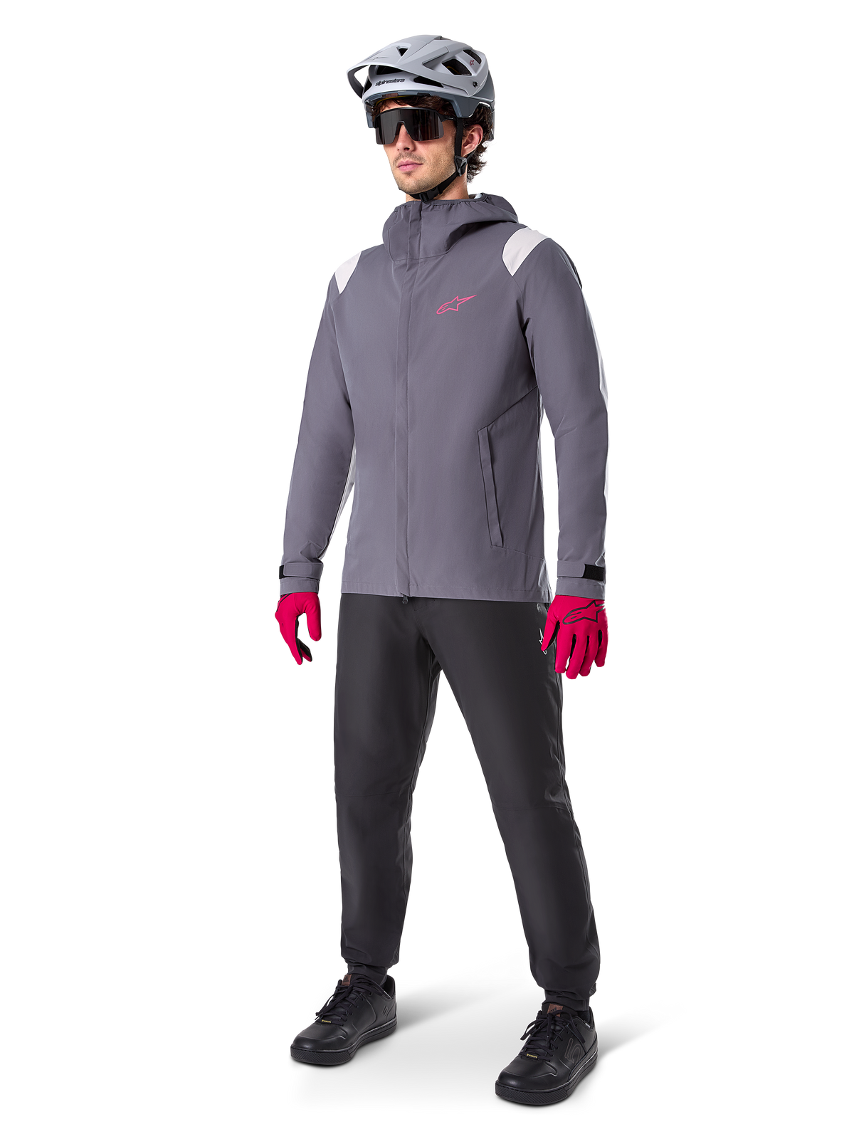 Mannelijk model draagt Alpinestars A-Dura Regenjas in Dark Titanium, grijze jas met capuchon met roze logo op de borst en lichtgrijze schouderpanelen, gecombineerd met een grijze MTB-helm, donkere zonnebril, felroze handschoenen en zwarte broek voor off-road fietsen.
