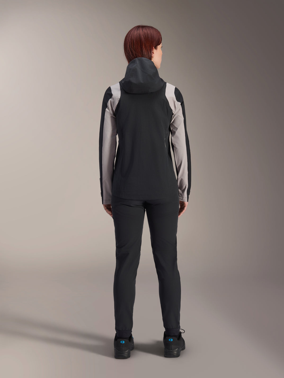 Alpinestars Stella A-Dura Regenjacke für Damen, Rückansicht einer Fahrerin, Schwarz mit hellgrauen Einsätzen an Ärmeln und Schultern, aufgesetzte Kapuze, kombiniert mit schwarzer Hose und Fahrradschuhen, technische Mountainbike-Regenausrüstung