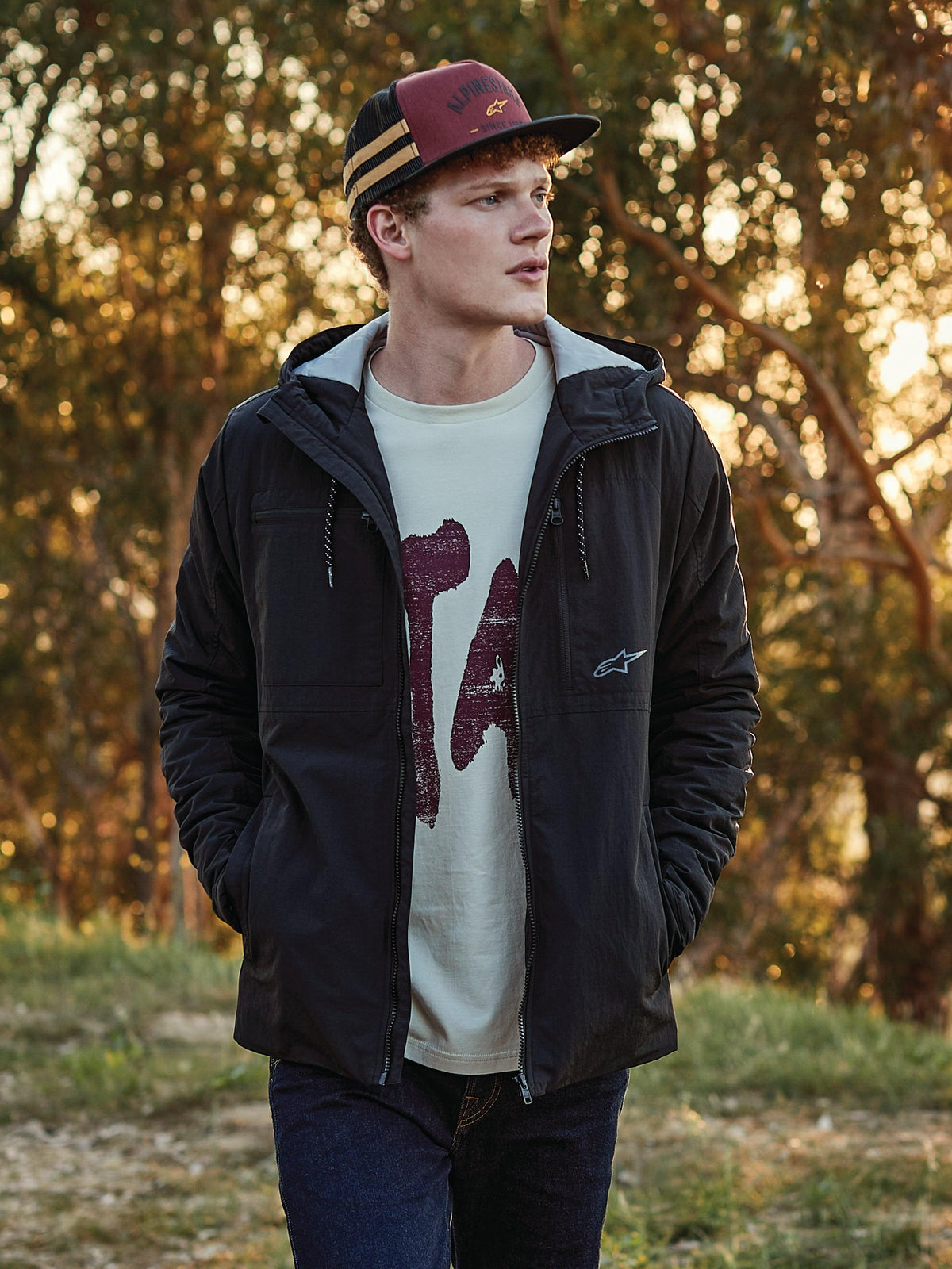 Alpinestars Backline Trucker Mütze in Ziegelrot, Schwarz und Senfgelb, getragen von einem Mann kombiniert mit einer schwarzen Kapuzenjacke und einem Grafik-T-Shirt, lässige vom Motorsport inspirierte Lifestyle-Streetwear, Außenaufnahme zur goldenen Stunde