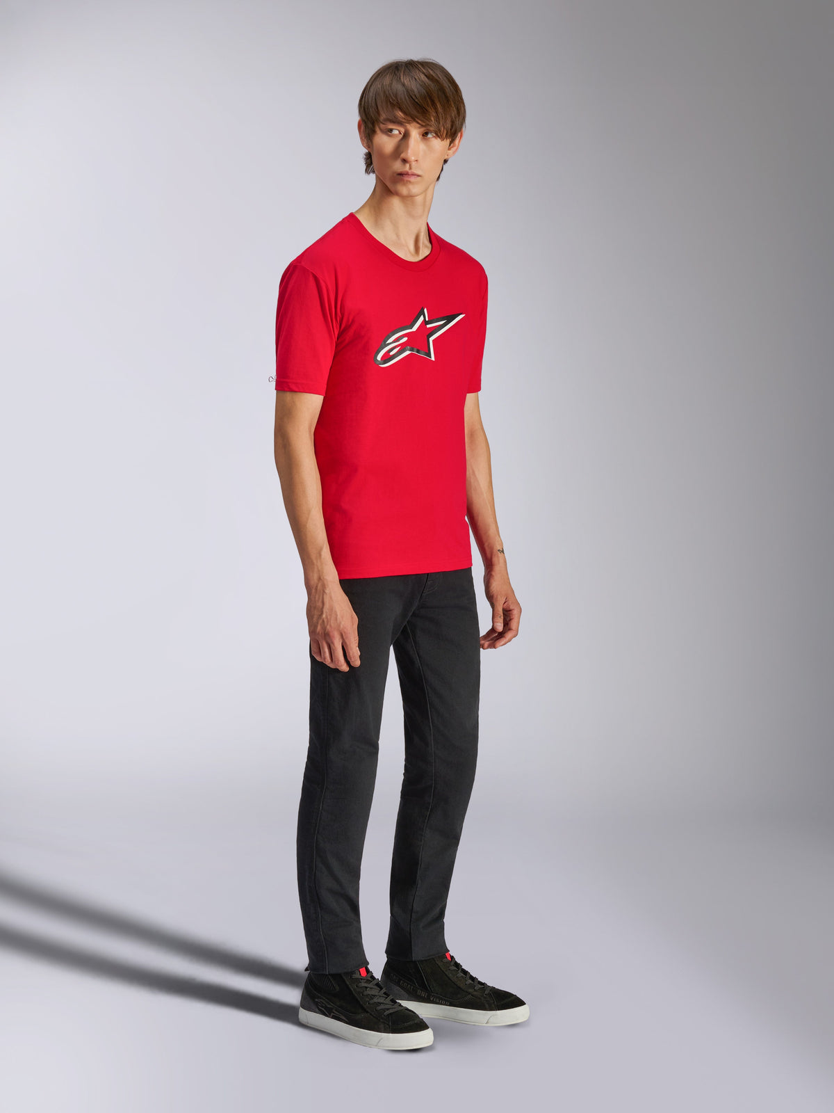 Alpinestars Ageless Shadow CSF Tee, rotes Kurzarm-T-Shirt, mit großem Ageless-Logo auf der Brust, getragen von einem Model mit schwarzer Hose und Sneakern, lässige Motorsport-Streetwear