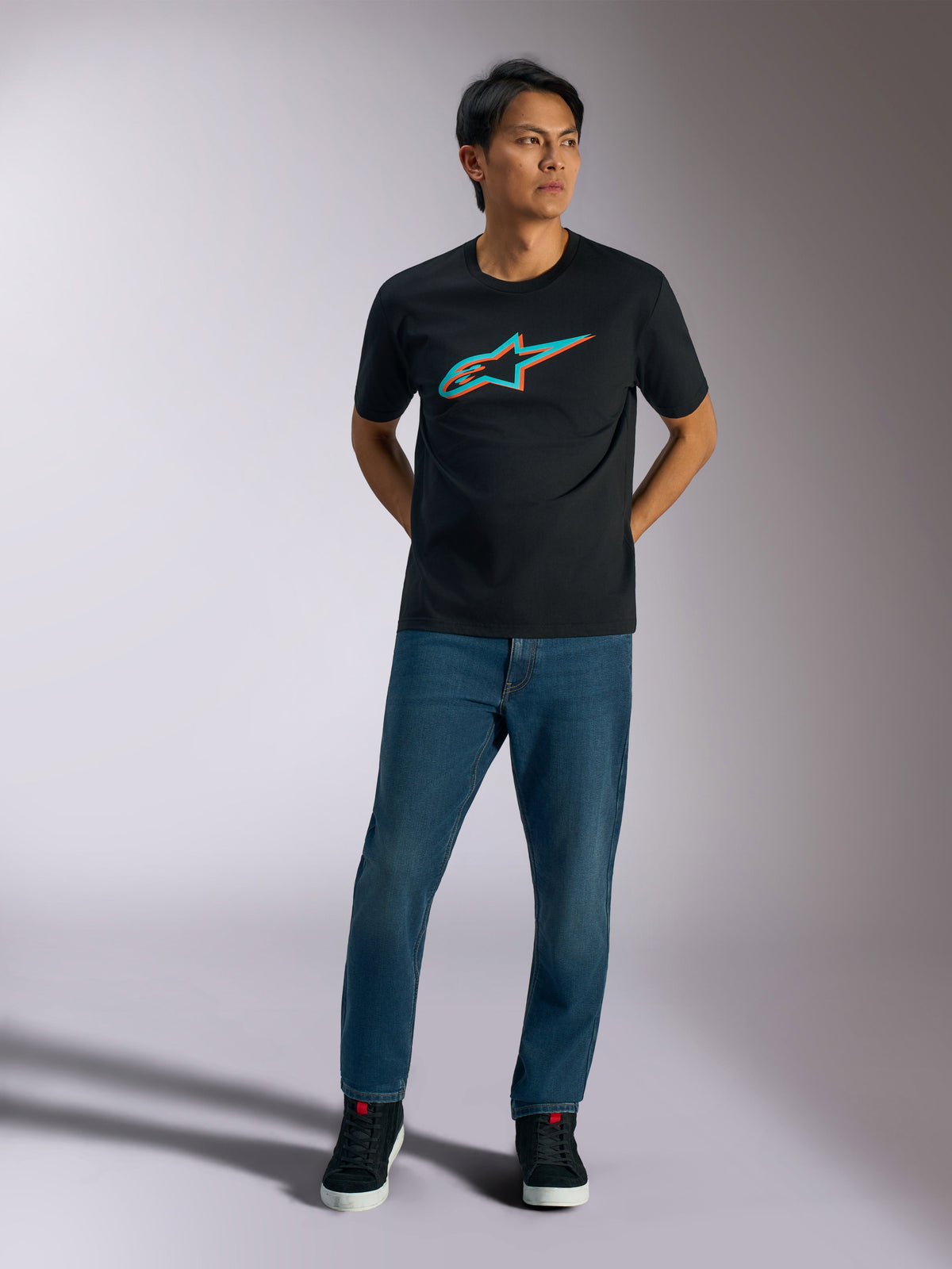Alpinestars Ageless Shadow CSF Tee, t-shirt met korte mouwen, zwart, turquoise en oranje Ageless shadow-logo op de borst, gedragen door model met blauwe jeans en zwarte schoenen, casual motorsport streetwear