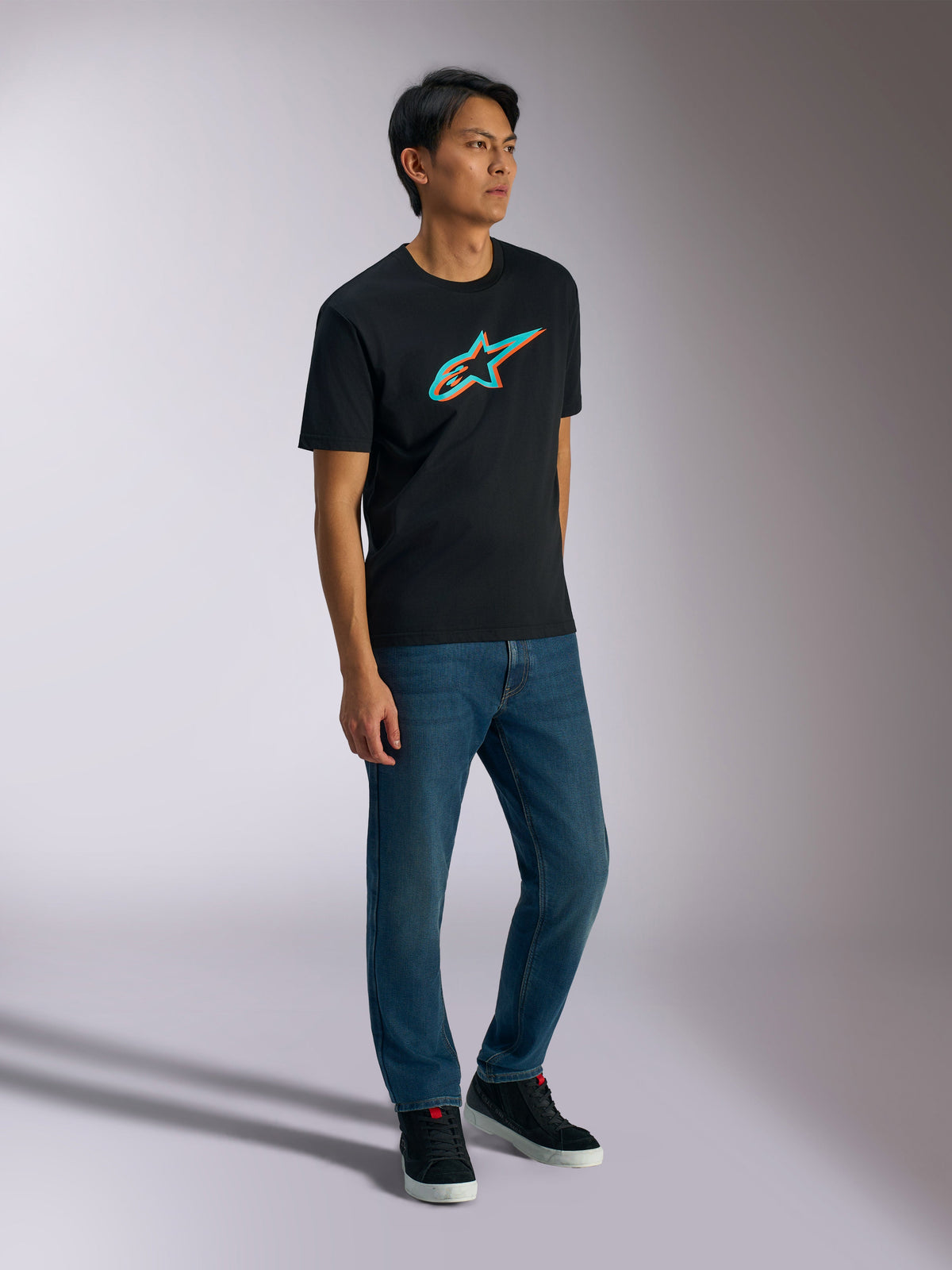 Alpinestars Ageless Shadow CSF Tee, T-shirt met korte mouwen, zwart met turquoise en oranje schaduwlogo, gedragen door mannelijk model met blauwe jeans en zwarte sneakers, casual op motorsport geïnspireerde streetwear