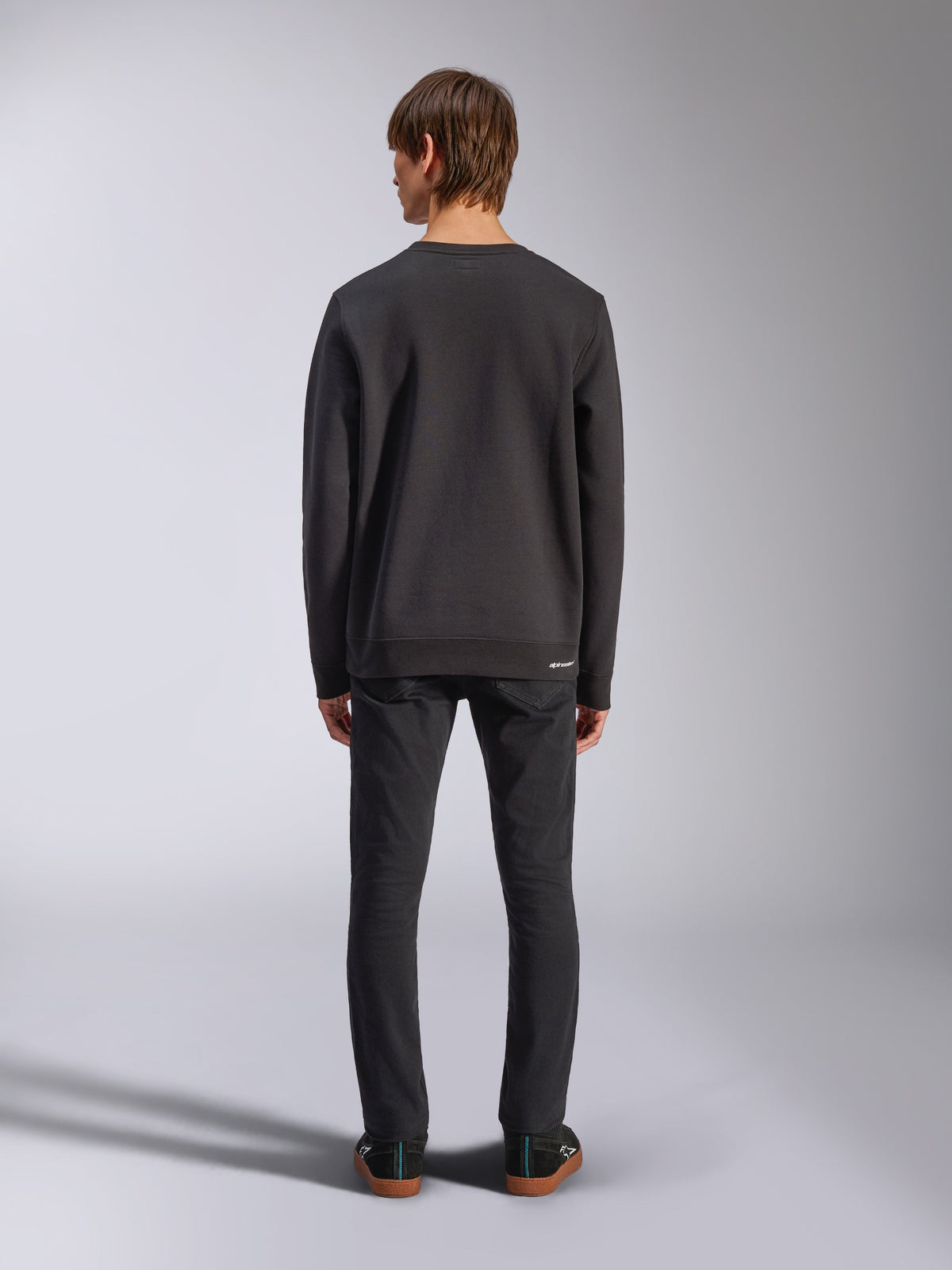 Alpinestars Positive Crew, zwarte pullover sweatshirt, achteraanzicht op mannelijk model, gedragen met zwarte jeans en casual sneakers, voorzien van geribde manchetten en zoom, op motorsport geïnspireerde streetwear