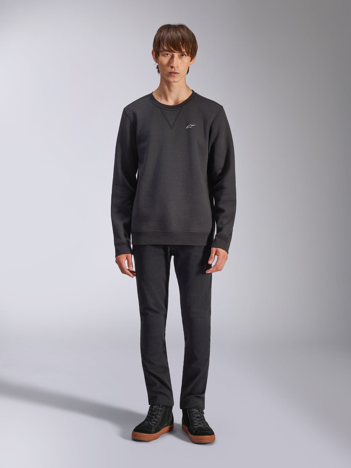 Alpinestars Positive Crew sweatshirt, casual zwarte sweater met ronde hals, klein wit logo op de borst, V-stiksel bij de halslijn, studio-opname van model in zwarte broek en sneakers, lifestylekleding geïnspireerd op de motorsport