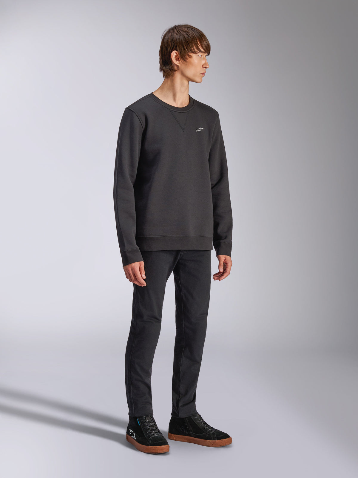 Alpinestars Positive Crew, pullover sweatshirt, zwart, klein wit Alpinestars-logo op de borst, geribbelde kraag met V-stikseldetail, geribbelde manchetten en zoom, gedragen door een model met zwarte broek en sneakers, casual motorsport-geïnspireerde streetwear