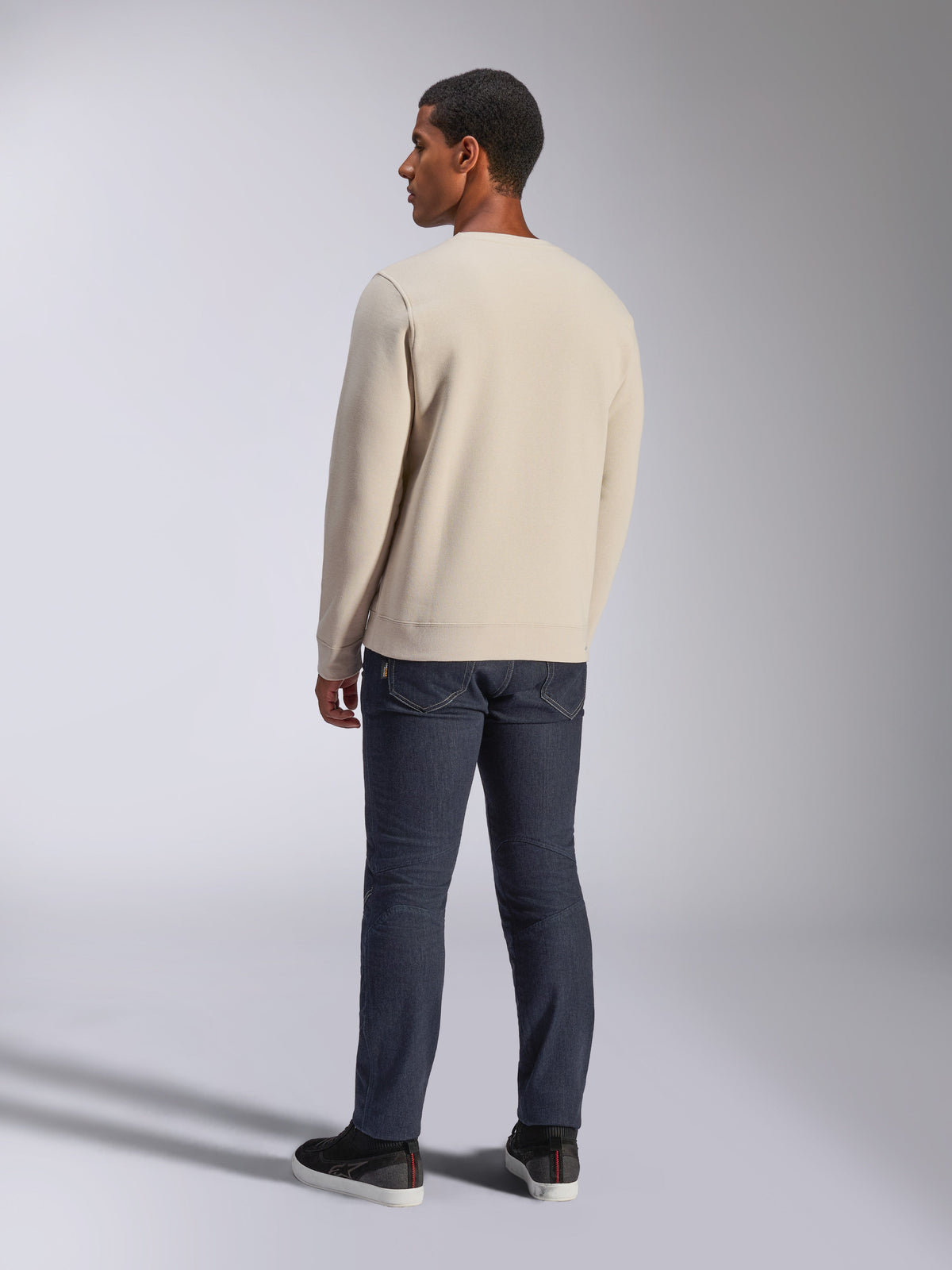 Alpinestars Positive Crew, Off White sweatshirt met ronde hals, achteraanzicht op model, gecombineerd met een donkere denim jeans en zwarte sneakers, casual motorsport streetwear, met geribde boorden en zoom
