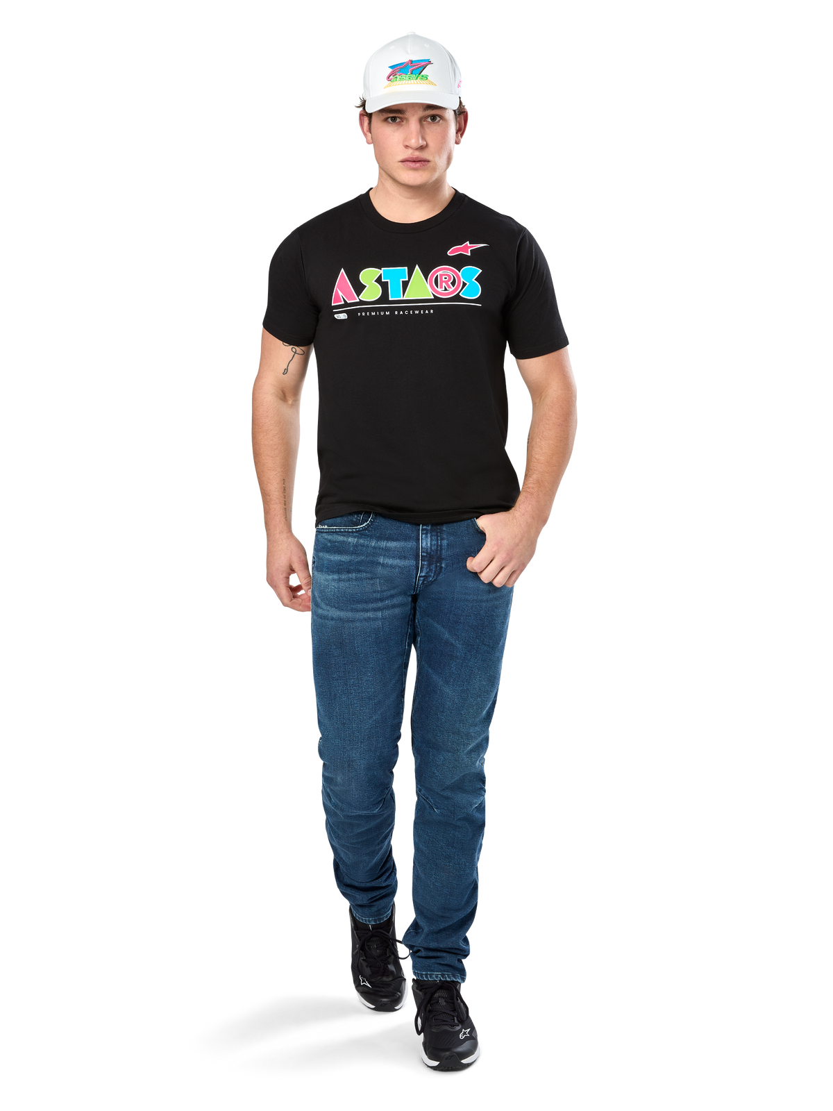 Modèle masculin portant le t-shirt Alpinestars Cereal CSF, t-shirt noir à manches courtes avec logo ASTARS géométrique coloré, associé à un jean bleu et une casquette blanche, style streetwear décontracté inspiré du sport automobile.
