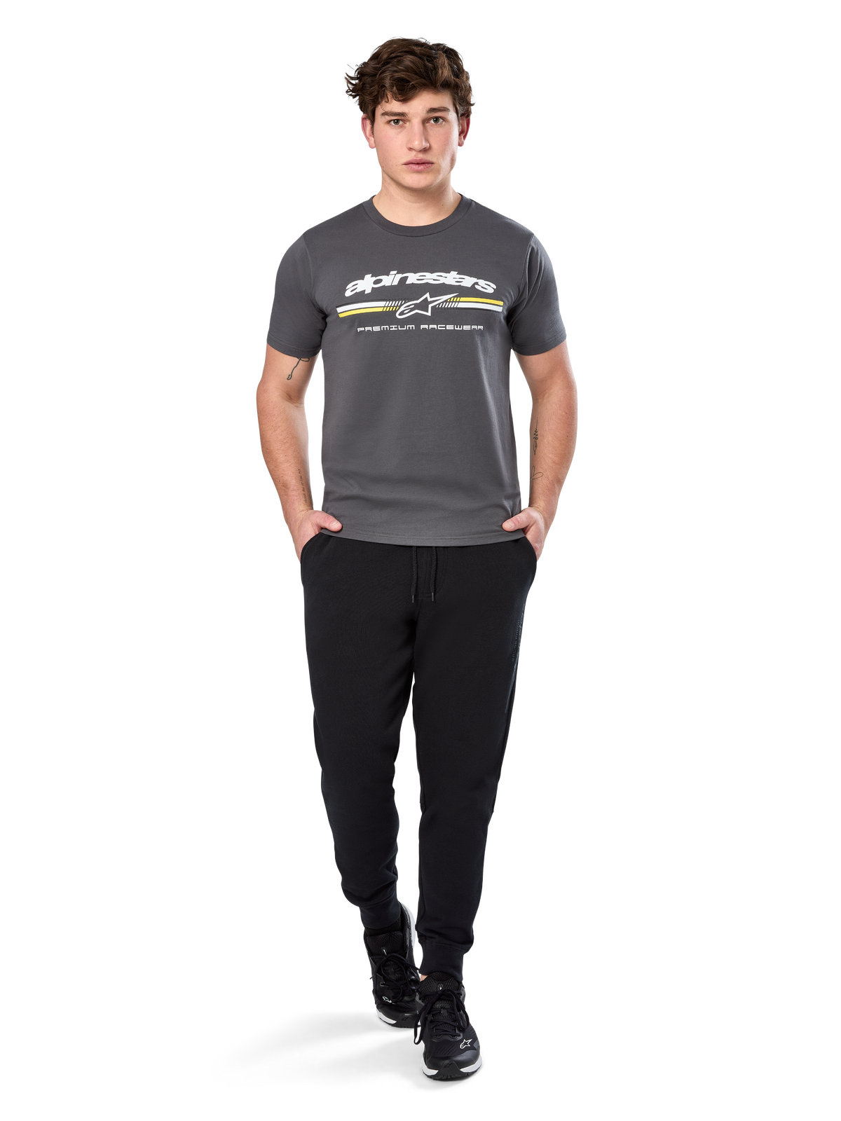 Homme portant le t-shirt à manches courtes Alpinestars Prevail CSF, gris anthracite, avec logo Alpinestars blanc et rayure tricolore aux accents jaunes, associé à un pantalon de jogging noir et des baskets, style motorsport décontracté