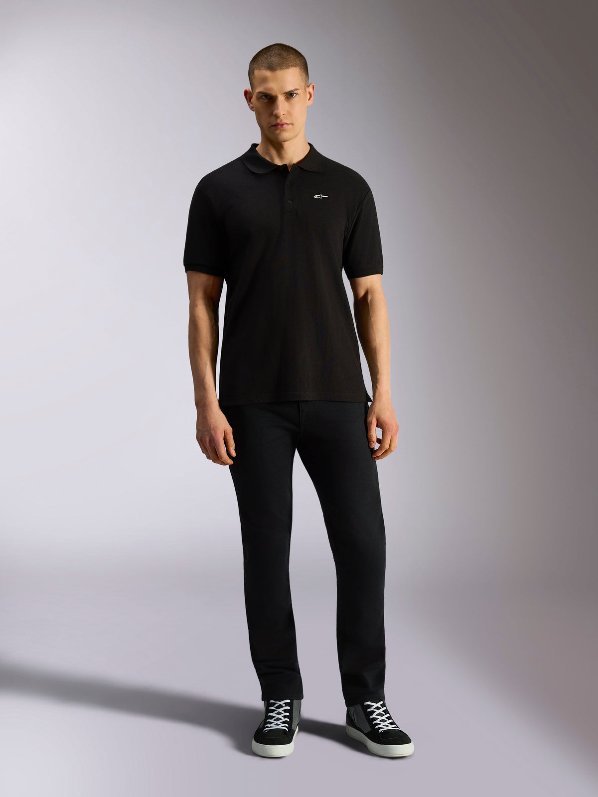 Alpinestars Endeavor Classic Polo, klassieke piqué polo met korte mouwen, zwart, klein wit Astars-logo op de borst, geribde kraag en manchetten, gedragen door model met zwarte broek en sneakers, casual motorsport streetwear