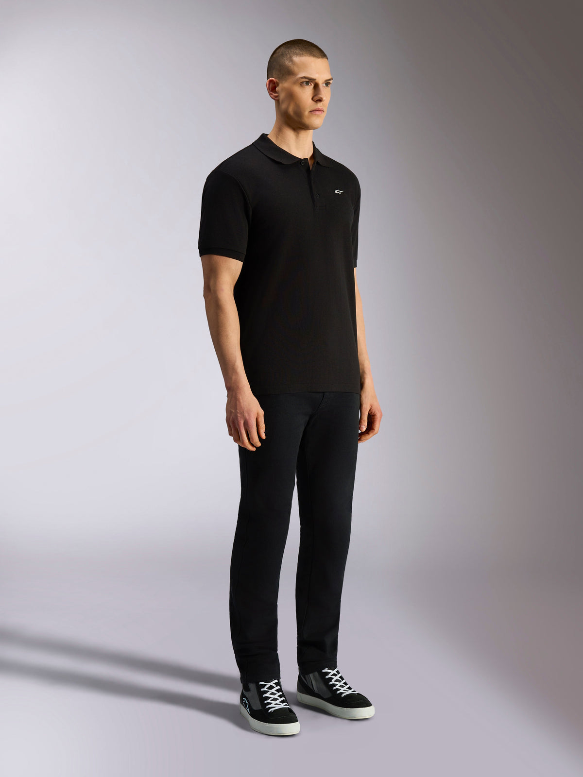 Alpinestars Endeavor Classic Polo, zwart poloshirt met korte mouwen, piqué gebreid met een knoopsluiting met drie knopen, klein wit Astars-logo op de linkerborst, gedragen met een zwarte broek en sneakers, casual motorsport streetwear