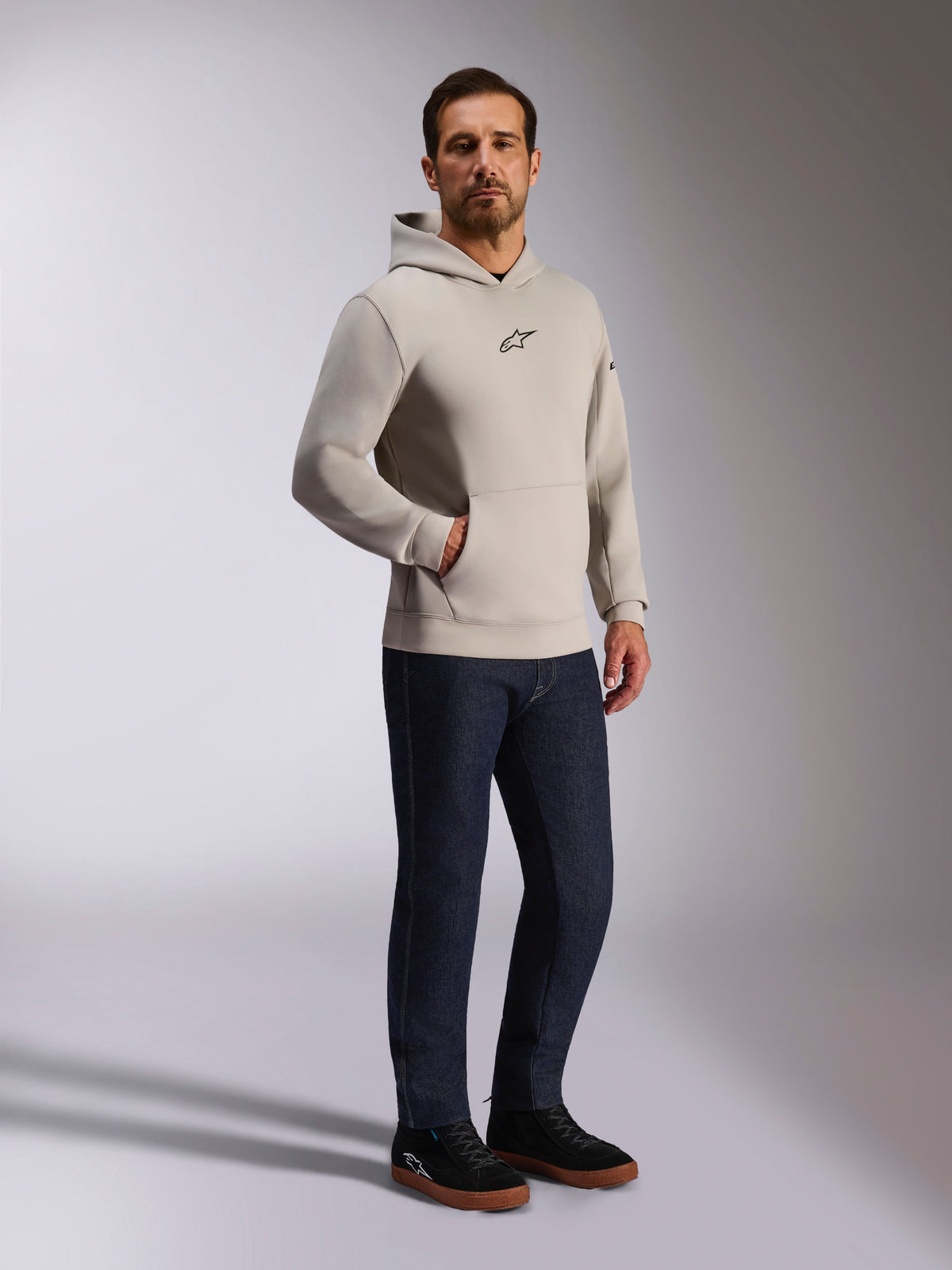 Alpinestars Insight Kapuzenpullover, lässige Streetwear, Zementgrau mit schwarzem Astars-Logo auf der Brust, Pullover-Design mit Kängurutasche, getragen mit dunkelblauer Jeans und schwarzen Sneakern, moderner, vom Motorsport inspirierter Stil