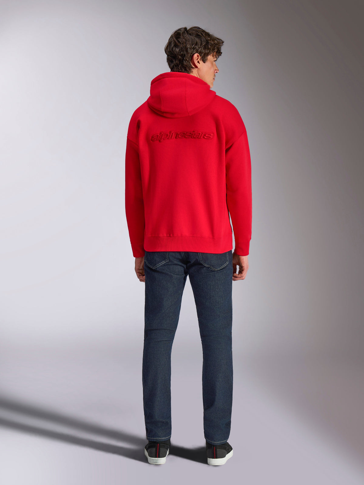 Alpinestars Emergent Oversized Hoodie, Rot, Rückansicht an einem männlichen Model, mit Ton-in-Ton 3D-besticktem Alpinestars-Wortlogo, kombiniert mit dunkelblauen Jeans, lässige motorsport-inspirierte Streetwear