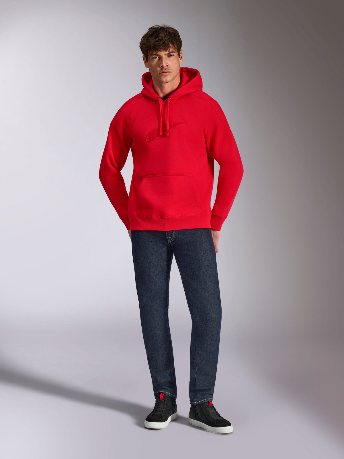Alpinestars Emergent Oversized Hoodie, Rot, mit tonaler 3D-Logo-Stickerei auf der Brust, Kängurutasche und Kapuze mit Kordelzug, getragen von einem Model mit dunkelblauer Jeans und schwarzen Sneakern, lässige Lifestyle-Streetwear