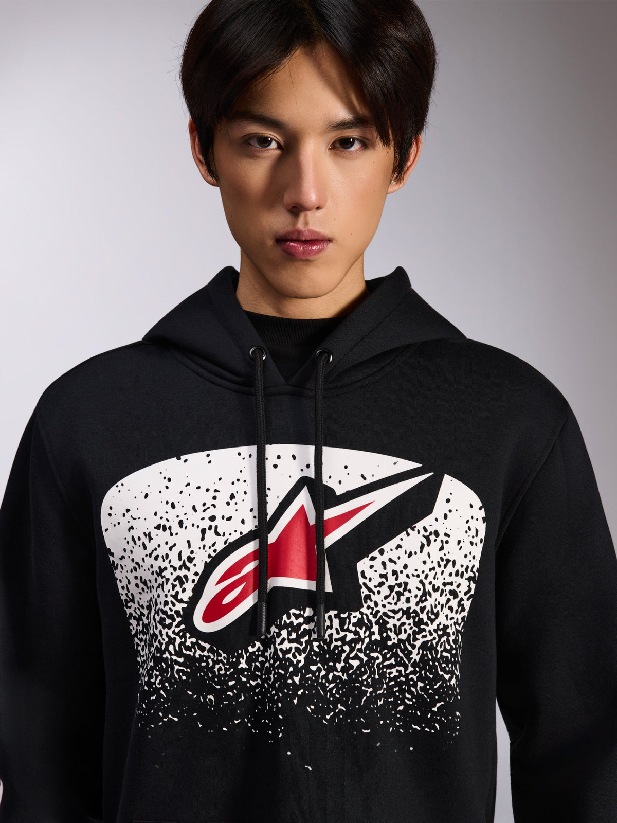 Alpinestars Obsessed Hoodie, sweat à capuche en polaire noire, logo Astars rouge et blanc dans un graphisme en dégradé MX pointillé blanc sur la poitrine, porté par un mannequin, streetwear décontracté avec capuche à cordon de serrage ajustable