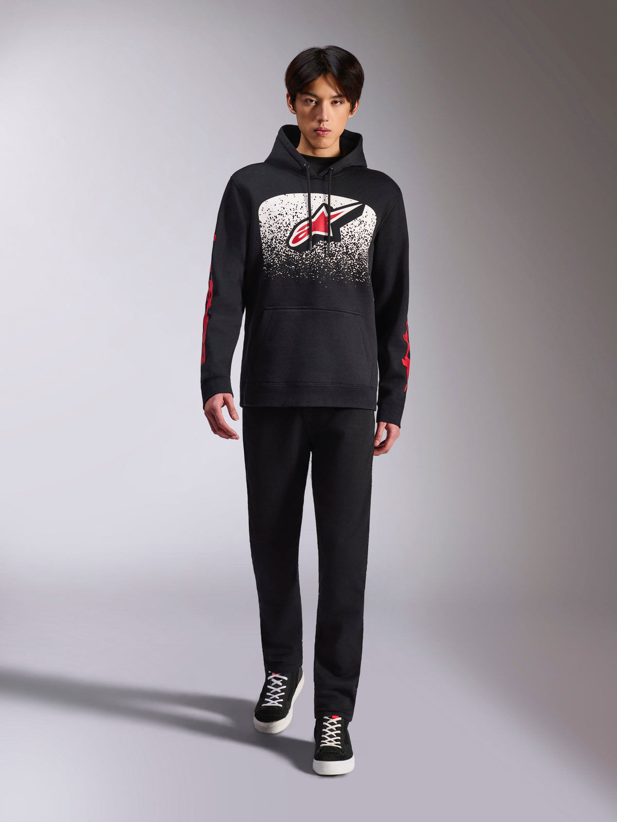 Sweat à capuche Alpinestars Obsessed, sweat à capuche noir à enfiler, grand logo Astars blanc et rouge avec dégradé MX, logos Alpinestars Word rouges sur les manches, porté par un mannequin dans une tenue décontractée entièrement noire, vêtement lifestyle inspiré du sport automobile