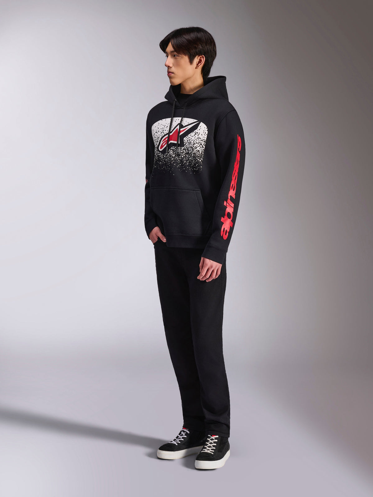 Sweat à capuche Alpinestars Obsessed, sweat en polaire noir à enfiler, avec un grand graphisme blanc moucheté sur la poitrine, logo Astars rouge et marquage rouge sur la manche, porté par un mannequin avec pantalon noir et baskets, style streetwear motorsport décontracté