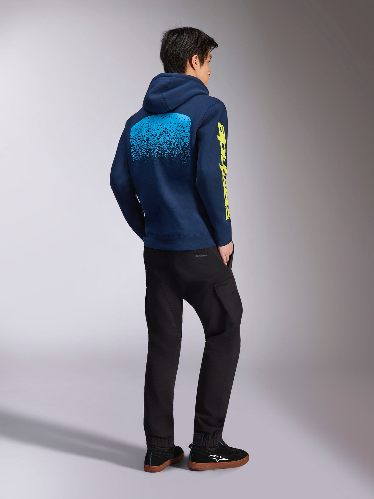 Alpinestars Obsessed Hoodie, dunkelblaues Freizeit-Sweatshirt, Rückansicht mit hellblauer MX-Farbverlaufsgrafik und gelbem Alpinestars-Logo am Ärmel, getragen mit schwarzer Hose und Sneakern