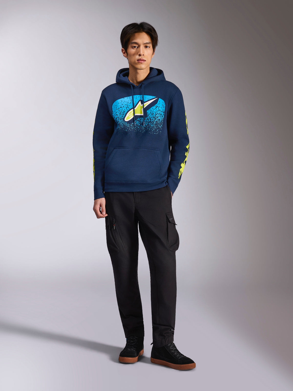 Alpinestars Obsessed Kapuzenpullover, dunkelblauer Hoodie, Astars-Logo vorne mit blauem MX-Farbverlauf und limettengelbem Stern, limettengelbe Alpinestars-Schriftzüge auf den Ärmeln, getragen von einem Model mit schwarzer Cargohose und schwarzen Sneakern, lässige Motorsport-inspirierte Streetwear