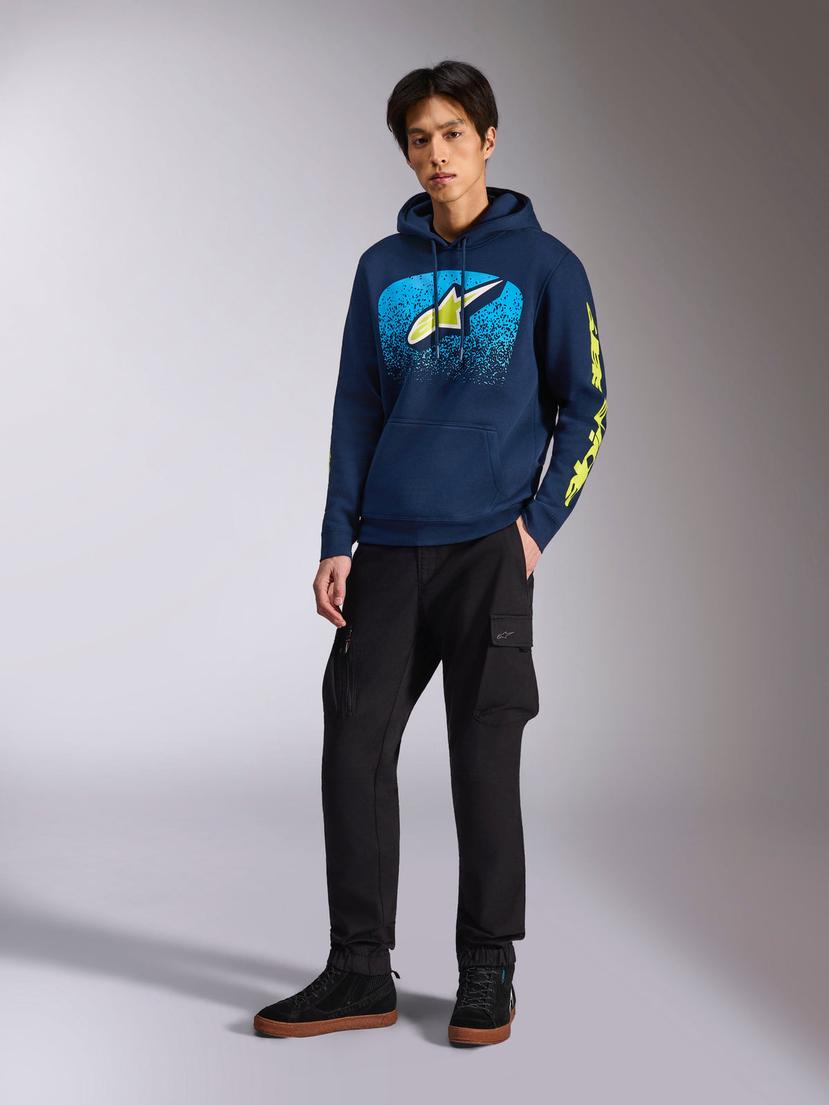 Alpinestars Obsessed Hoodie in Navy, lässiger Fleece-Kapuzenpullover mit hellblauer und limettengelber Grafik auf der Vorderseite, limettengelbes Alpinestars-Branding am Ärmel, getragen von einem Modell mit schwarzer Cargohose und schwarzen Sneakern, Motorsport-inspirierte Streetwear