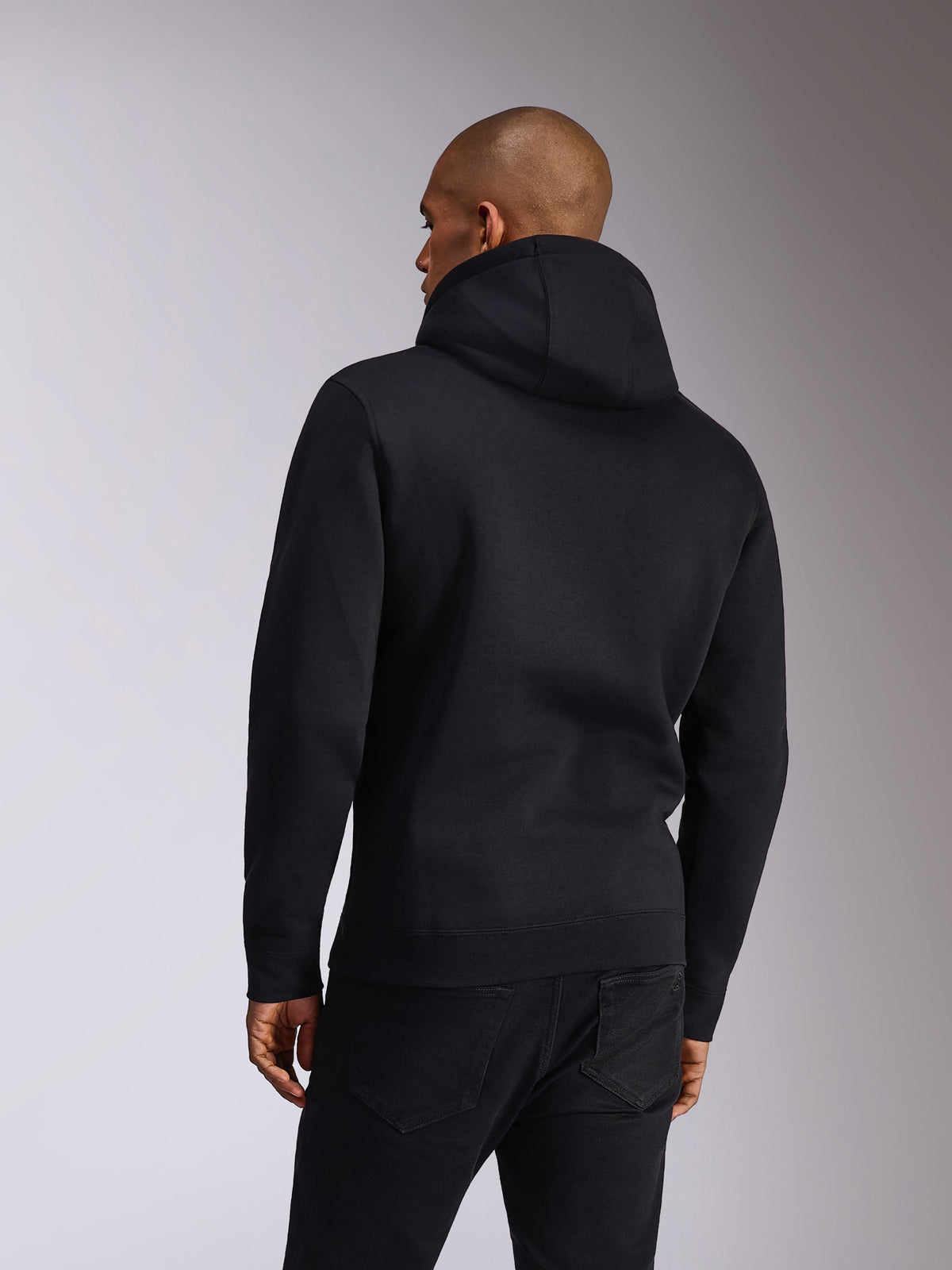 Alpinestars Ellipsoid Hoodie, Schwarz, Rückansicht einer Person, die einen Fleece-Kapuzenpullover trägt, lange Ärmel mit Rippbündchen an Ärmeln und Saum, kombiniert mit schwarzen Jeans, lässige Motorsport-inspirierte Sportbekleidung
