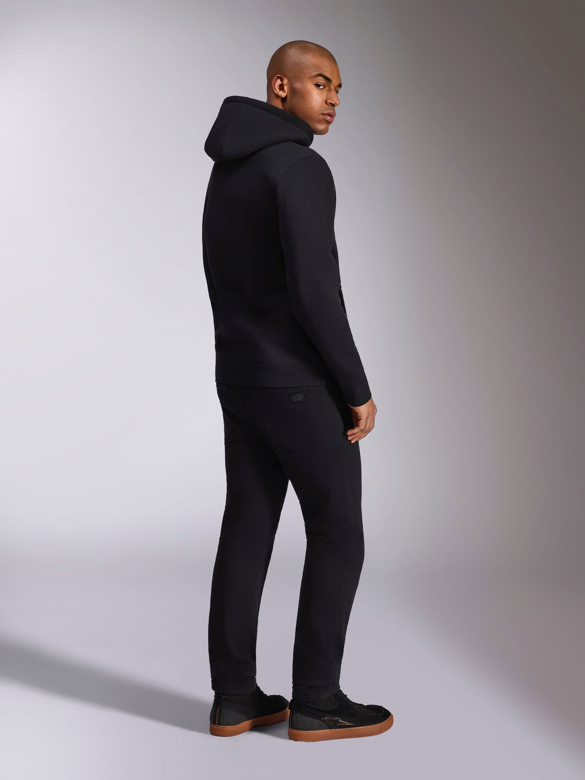 Alpinestars Ellipsoid Hoodie, schwarze Freizeit-Streetwear, Rückansicht an einem männlichen Model, Fleece-Kapuzenpullover mit Rippbündchen und Saum, getragen mit schwarzer Hose und Sneakern, modernes, vom Motorsport inspiriertes Design