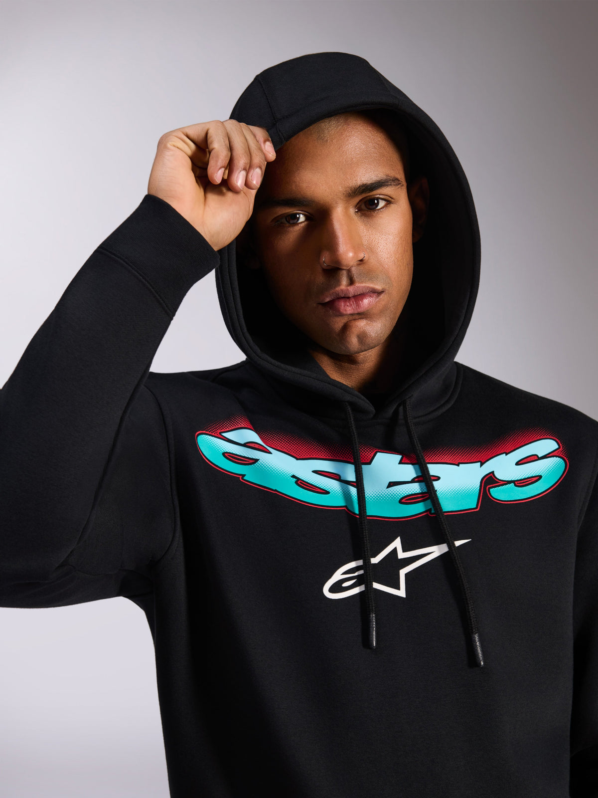 Alpinestars Ellipsoid Hoodie, schwarzer Fleece-Kapuzenpullover, mit stilisiertem Astars-Schriftzug im Farbverlauf von Blaugrün zu Rot und weißem Logo auf der Brust, getragen von einem Model mit aufgesetzter Kapuze, lässige vom Motorsport inspirierte Streetwear
