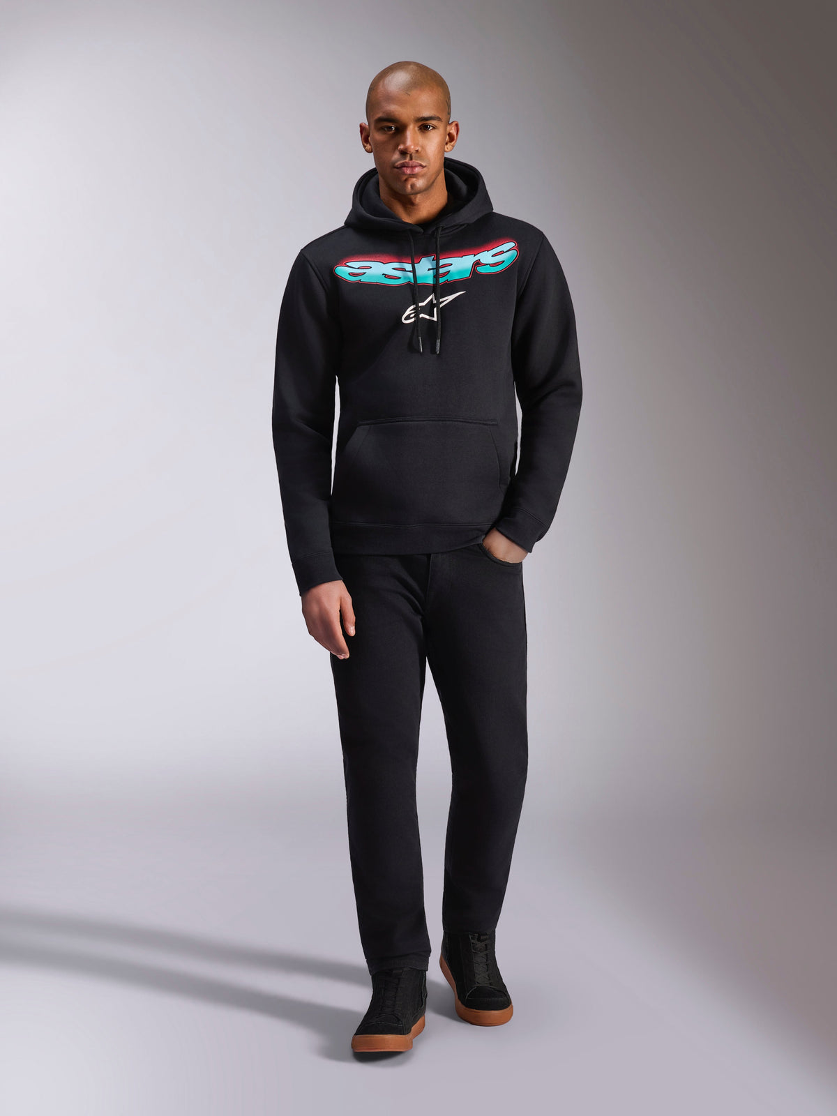 Alpinestars Ellipsoid Hoodie, Schwarz, Fleece-Kapuzenpullover mit geschwungenem Astars-Schriftzug in einem rot-türkisen Farbverlauf und weißem Stern-Logo, getragen von einem Model mit schwarzer Hose und Sneakern, lässige Motorsport-Streetwear