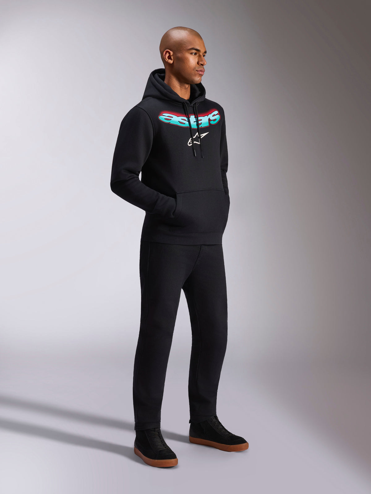 Alpinestars Ellipsoid Hoodie, schwarzer Fleece-Kapuzenpullover mit einem geschwungenen Astars-Schriftzug in türkis-rotem Farbverlauf und weißem Stern-Logo, getragen von einem Modell mit schwarzer Hose, lässige, vom Motorsport inspirierte Streetwear