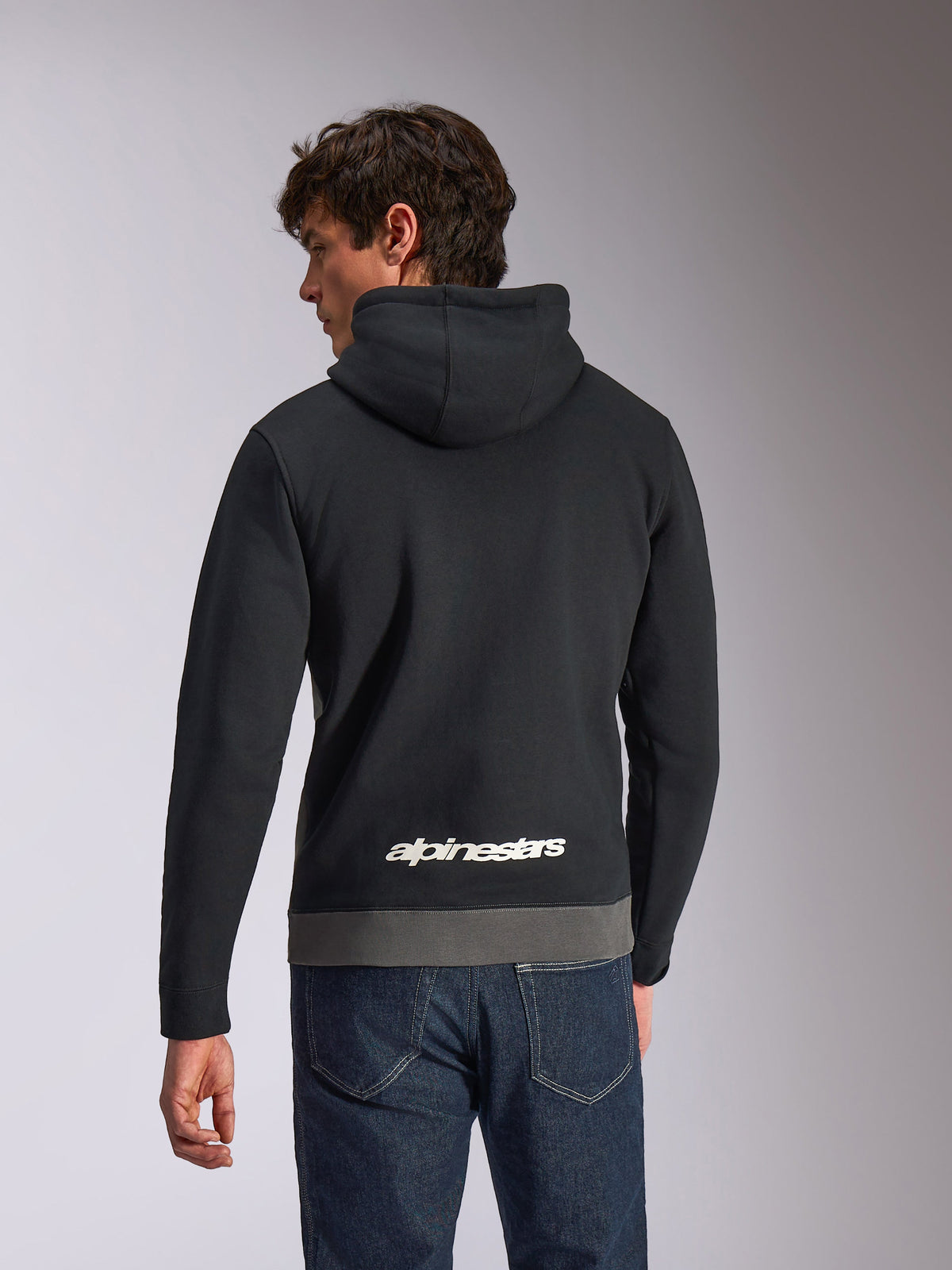 Alpinestars Parallel Hoodie, lässiger Sportswear-Kapuzenpullover, Schwarz und Gunmetal, Rückansicht mit weißem Alpinestars-Logo im unteren Rückenbereich, Color-Block-Design mit grauem Saum, kombiniert mit dunkler Jeanshose