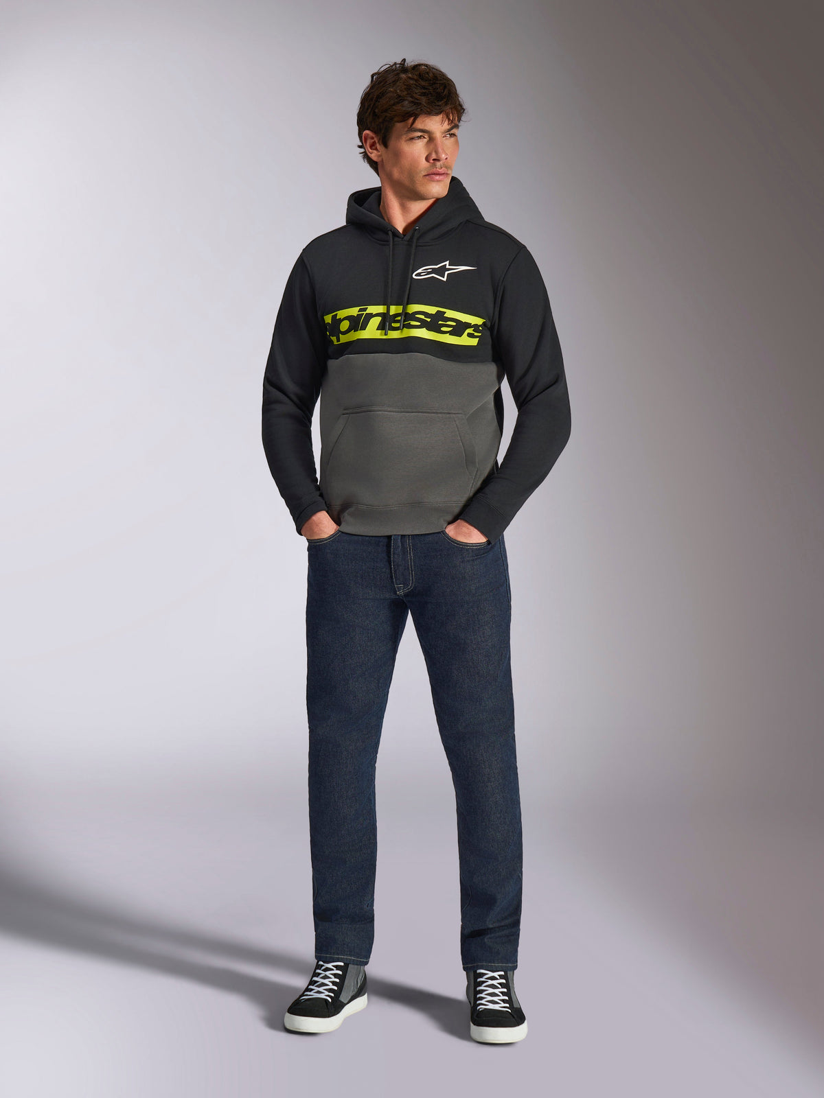 Alpinestars Parallel Hoodie, Color-Block-Kapuzenpullover in Schwarz und Gunmetal-Grau, leuchtend gelbes Alpinestars-Wortlogo und weißes Stern-Logo auf der Brust, getragen von einem männlichen Model mit dunkelblauer Jeans und schwarzen Sneakern, lässige Motorsport-Streetwear