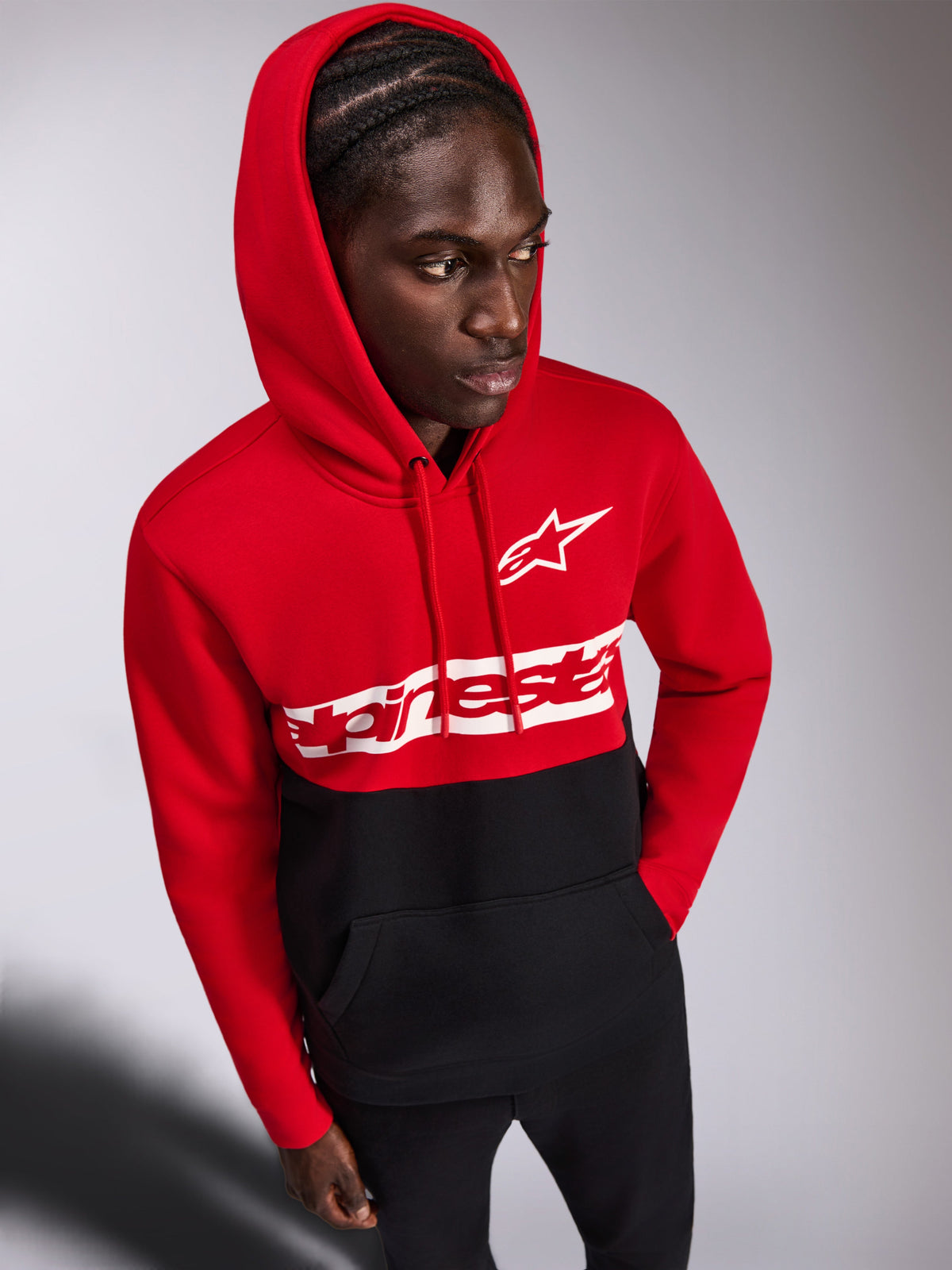 Alpinestars Parallel Hoodie, casual fleece pullover, rood en zwart color-blocked ontwerp, wit Alpinestars woordlogo en Astars-logo op de borst, gedragen door een man met de capuchon op en een zwarte broek, motorsport-geïnspireerde streetwear