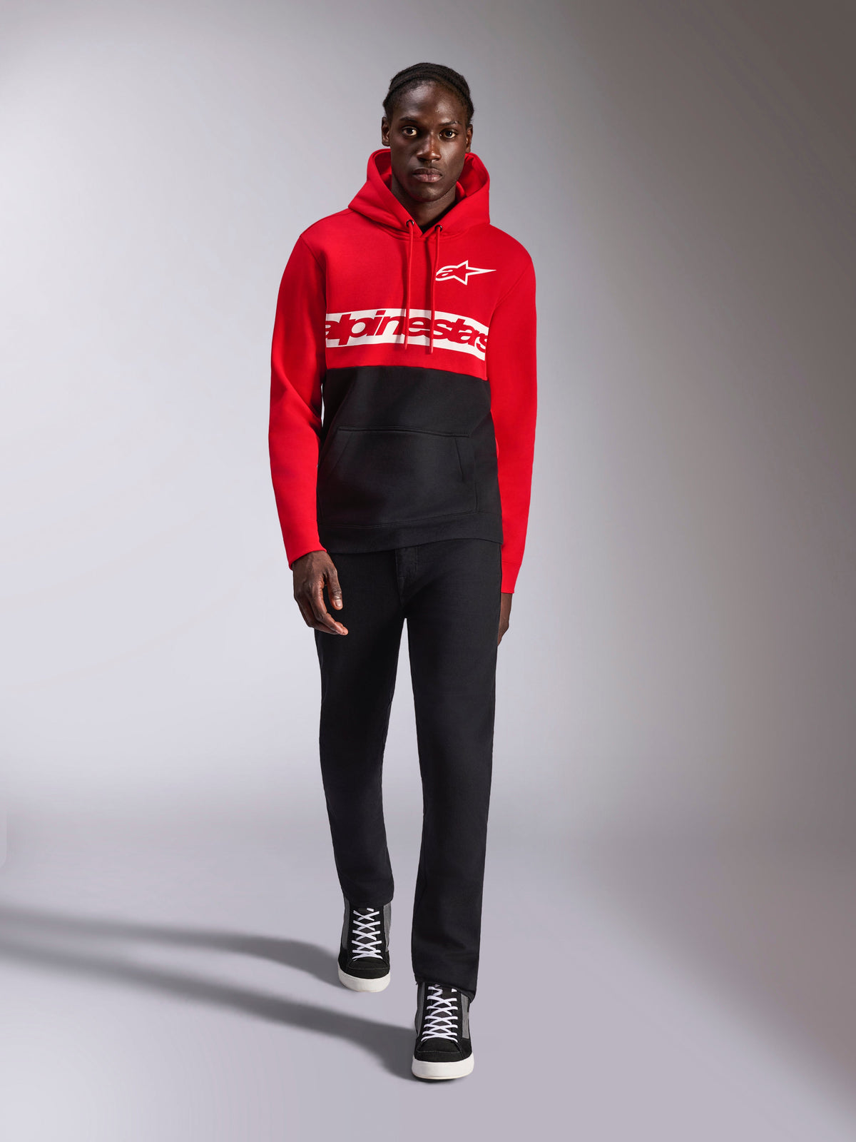 Alpinestars Parallel Hoodie, color-blocked fleece hoodie, rood en zwart, met wit Alpinestars-woordlogo op de borst en Astars-logo op de linkerborst, gedragen door een model met zwarte broek en hoge sneakers, casual motorsport streetwear