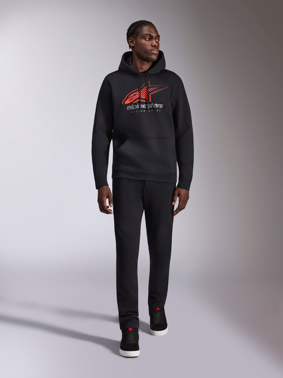 Alpinestars Pulse Hoodie, zwarte casual streetwear hoodie, rood en grijs Astars combo-logo met speed line fade op de borst, capuchon met trekkoord, kangoeroezak aan de voorkant, lange mouwen, gecombineerd met een zwarte broek en sneakers, op de motorsport geïnspireerd design