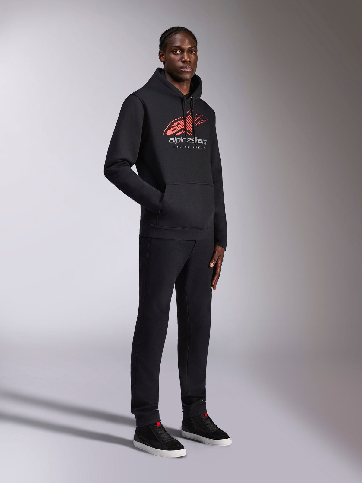 Alpinestars Pulse Hoodie, zwarte pullover hoodie, voorzien van rood en wit Astars-logo met vervagende speedlines en 'Racing Ahead' tekst, gedragen met bijpassende zwarte broek en sneakers, casual streetwear geïnspireerd op de motorsport.
