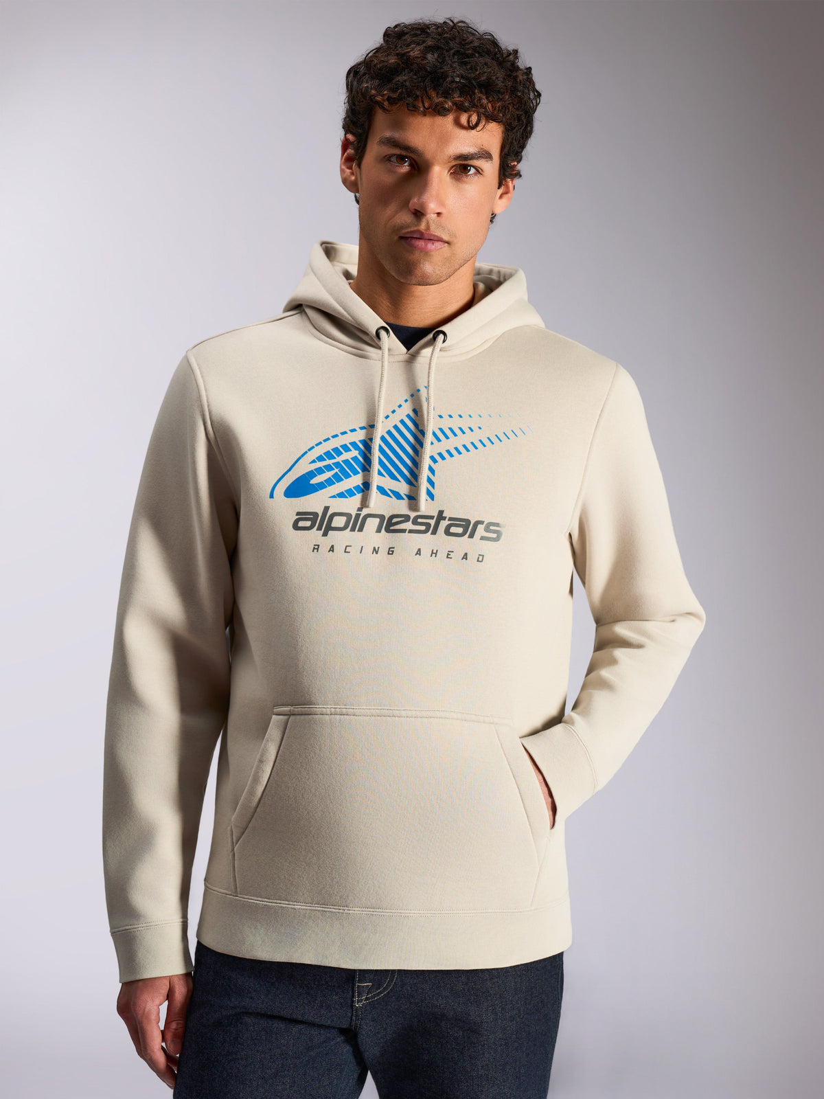 Sweat à capuche Alpinestars Pulse, streetwear décontracté, couleur sable, logo Astars Combo bleu et gris à l'avant avec dégradé de lignes de vitesse et texte 'Racing Ahead', capuche à cordon, poche kangourou, porté par un mannequin avec un jean foncé