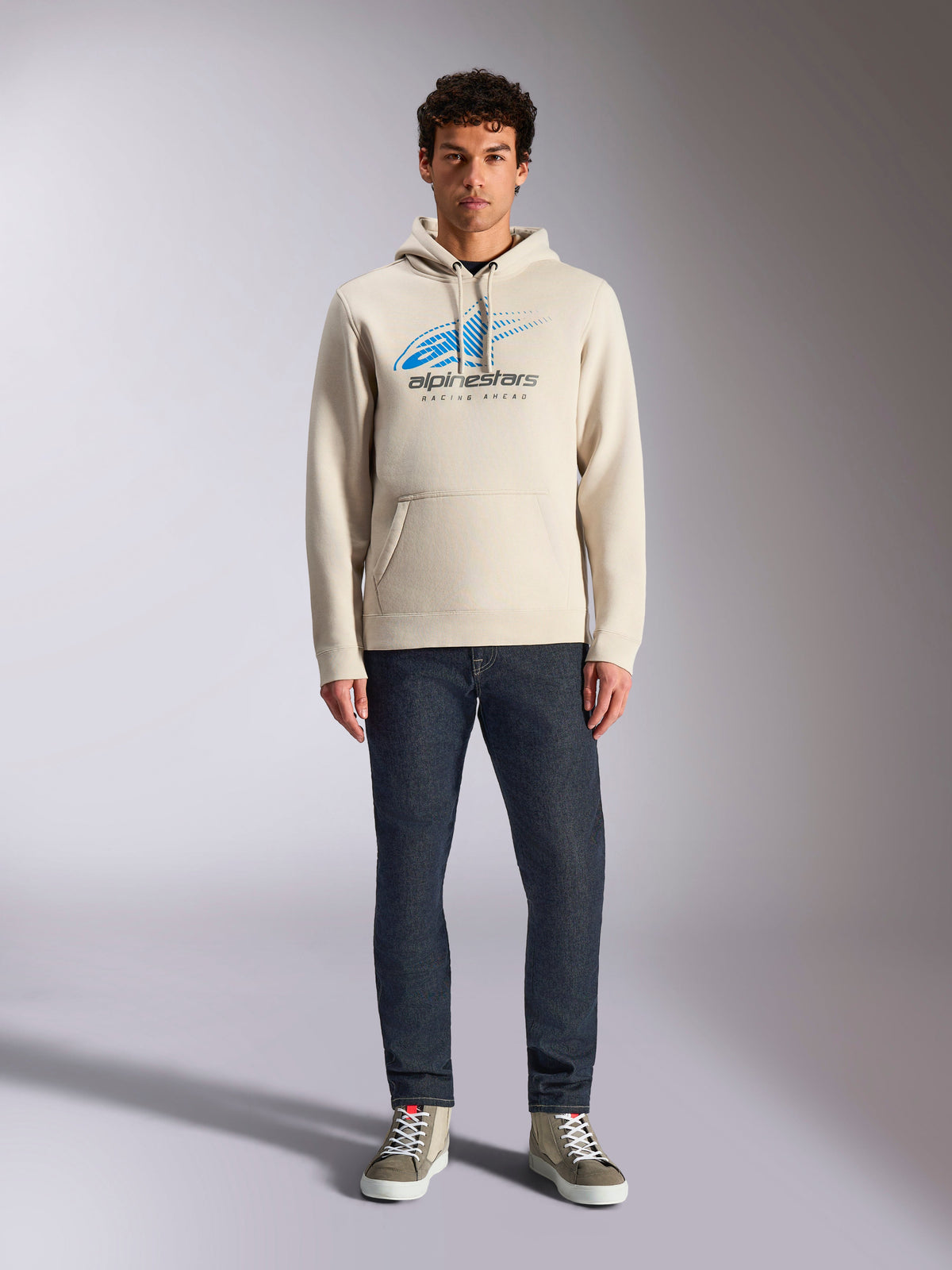 Sweat à capuche Alpinestars Pulse, blanc cassé, pull à manches longues avec capuche à cordon, logo Astars Combo bleu et noir avec dégradé de lignes de vitesse, porté avec un jean foncé et des baskets, style streetwear motorsport décontracté