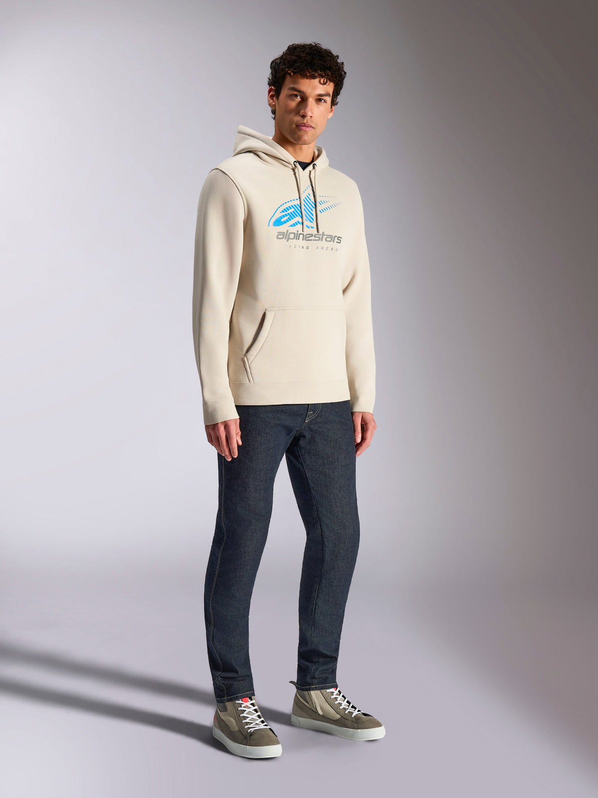 Sweat à capuche Alpinestars Pulse, style streetwear décontracté blanc cassé, sweat en polaire avec logo Astars bleu et gris et dégradé de lignes de vitesse, poche kangourou, porté par un mannequin avec un jean en denim foncé et des baskets montantes.