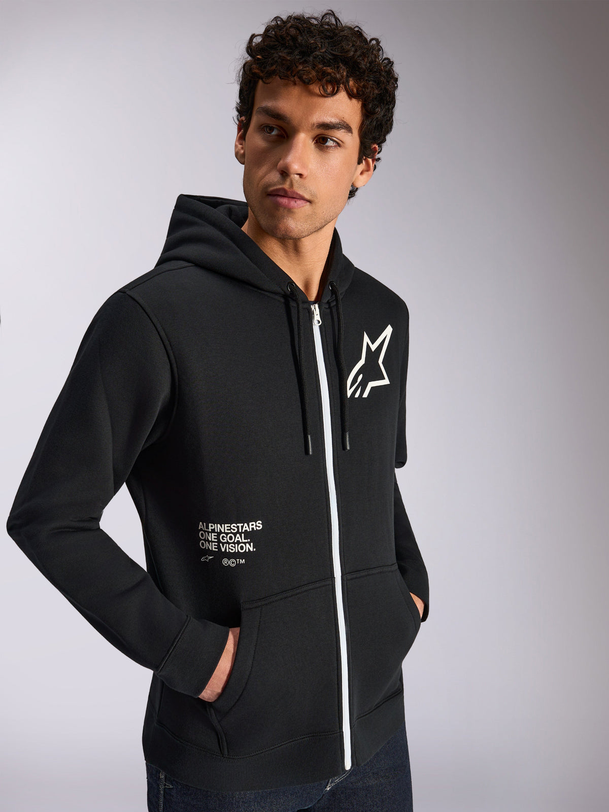Alpinestars Transverse Hoodie, casual sportswear, black with white zipper detail, drawstring hood, Alpinestars star logo on chest, 'One Goal One Vision' graphic, worn by male model with dark curly hair