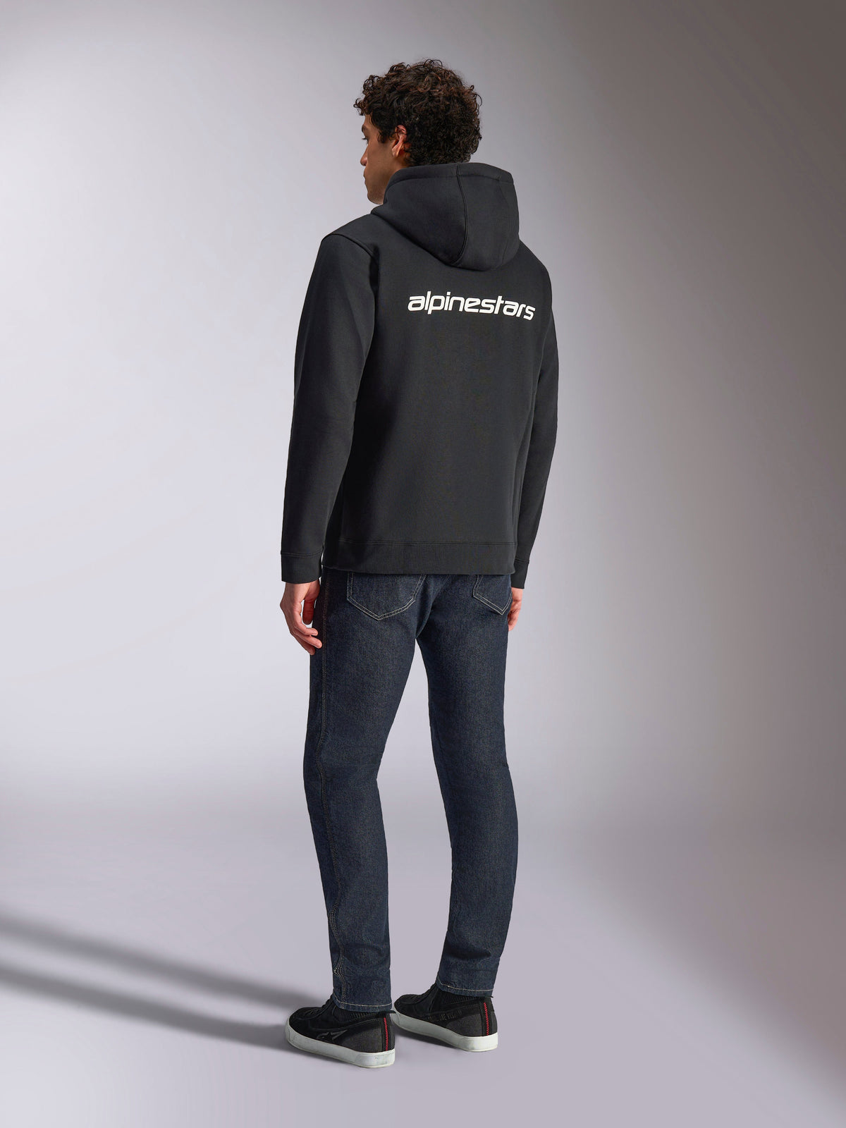 Alpinestars Transverse Hoodie, Black zip-front fleece, back view featuring a white Alpinestars word logo across the shoulders, worn with dark blue jeans and black sneakers, casual motorsport-inspired streetwear