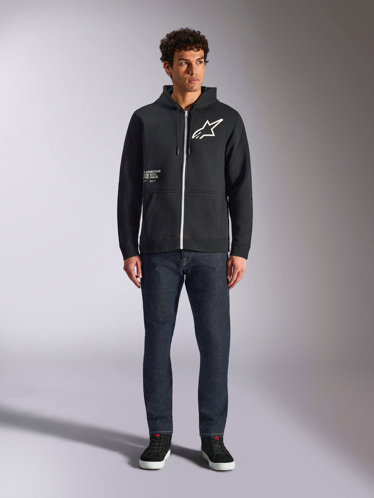 Alpinestars Transverse Hoodie, casual zip-front fleece, Black, white Astars logo on chest and 'One Goal' print on front, drawstring hood, paired with dark jeans and black sneakers, lifestyle motorsport streetwear