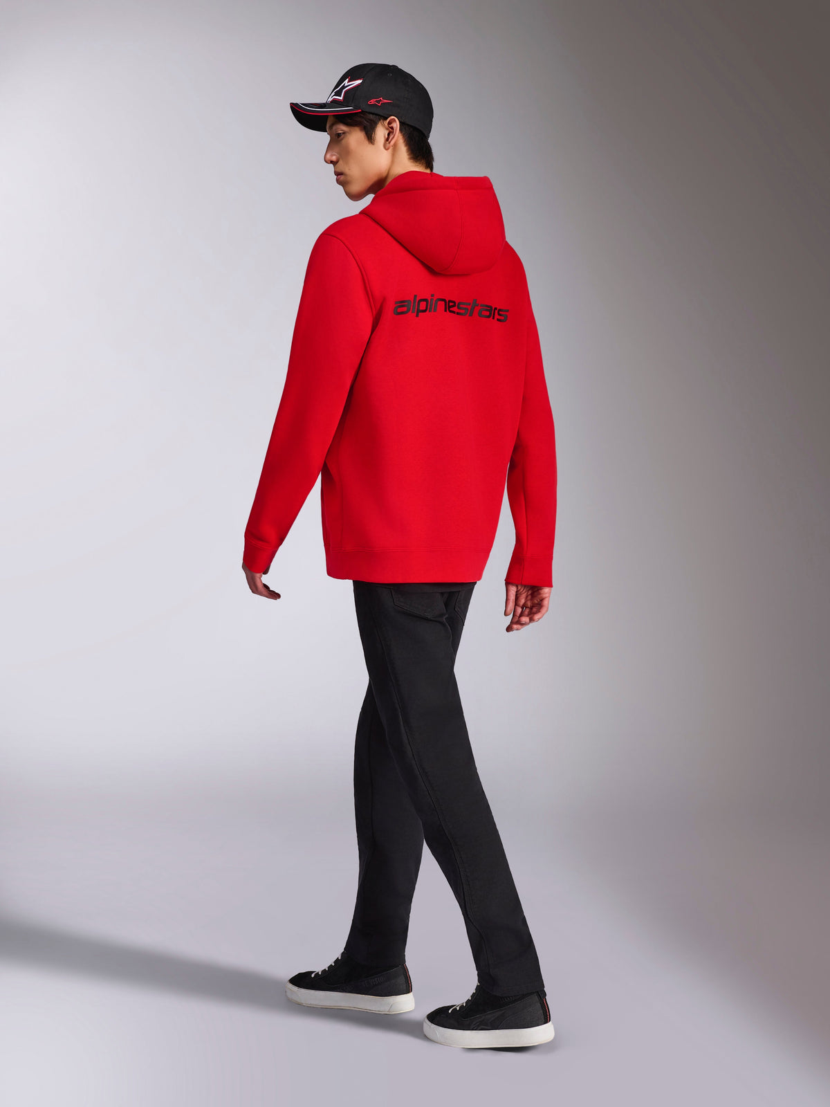Alpinestars Transverse Hoodie, Roter Fleece-Kapuzenpullover mit Reißverschluss, Rückansicht mit schwarzem Alpinestars-Wortlogo, getragen von einem Model mit schwarzer Hose und schwarzer Logo-Kappe, lässige, vom Motorsport inspirierte Streetwear