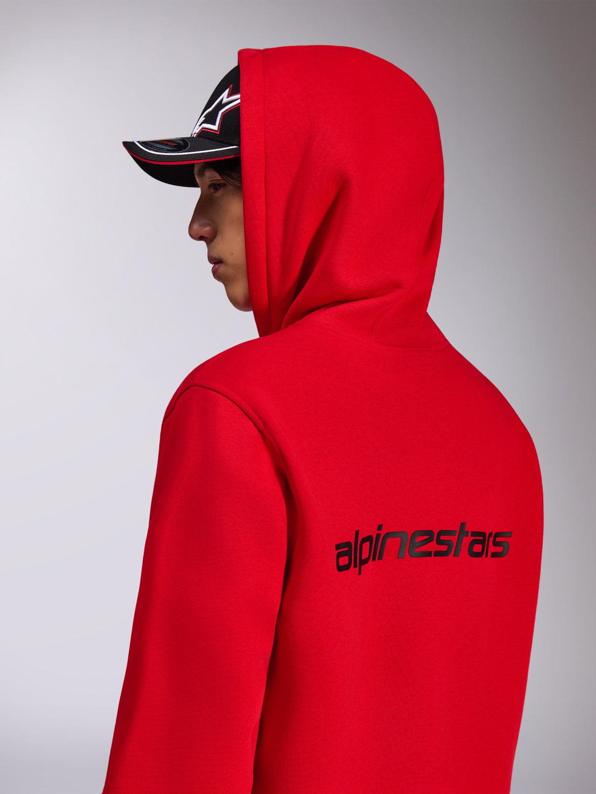 Alpinestars Transverse Hoodie, roter Kapuzenfleece, Seiten-Rückansicht mit schwarzem Alpinestars-Schriftzug auf dem Rücken, getragen mit hochgezogener Kapuze und einer schwarzen Kappe, lässige, vom Motorsport inspirierte Streetwear