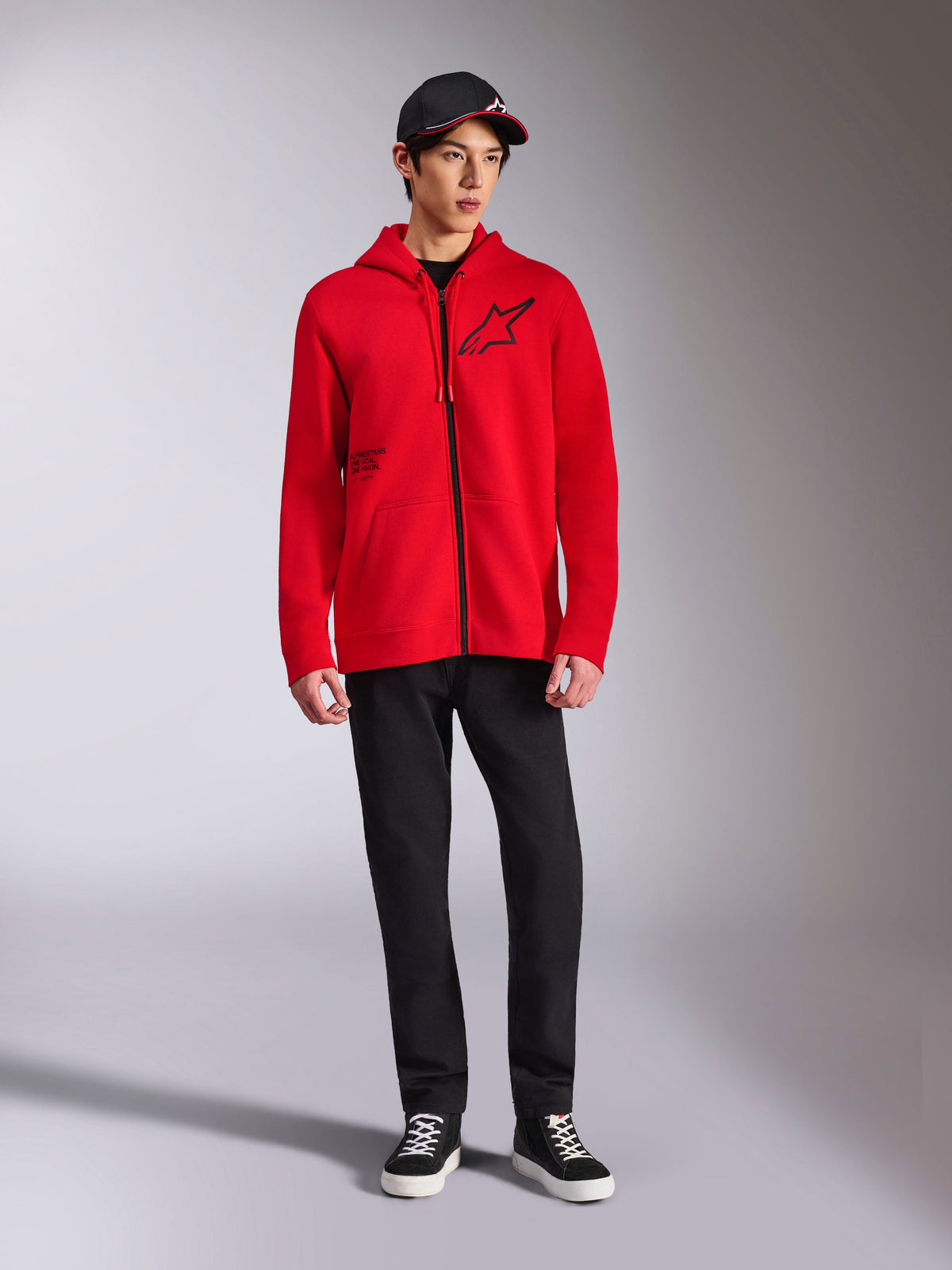 Alpinestars Transverse Hoodie, roter Fleece-Kapuzenpullover mit durchgehendem Reißverschluss, schwarzes Astars-Logo auf der Brust und One Goal-Druck an der Seite, getragen von einem männlichen Model mit schwarzer Hose und Kappe, lässige, vom Motorsport inspirierte Streetwear