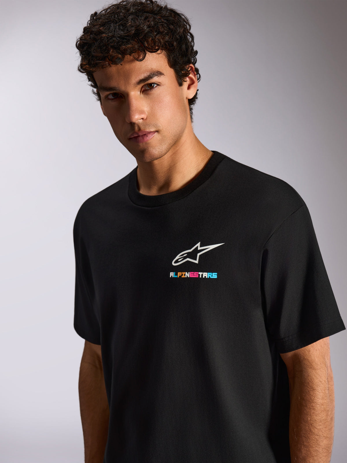 Un homme portant le T-shirt Alpinestars Wild Oversized CSF, t-shirt noir à manches courtes avec un logo Astars blanc et un marquage multicolore vif sur la poitrine, streetwear décontracté motorsport