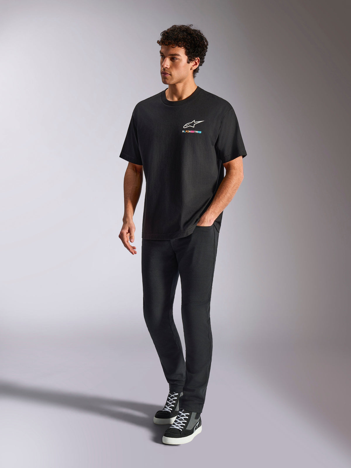 T-shirt Alpinestars Wild Oversized CSF, t-shirt à manches courtes, noir, logo Astars multicolore sur la poitrine, style streetwear décontracté porté par un mannequin avec un pantalon noir et des baskets montantes