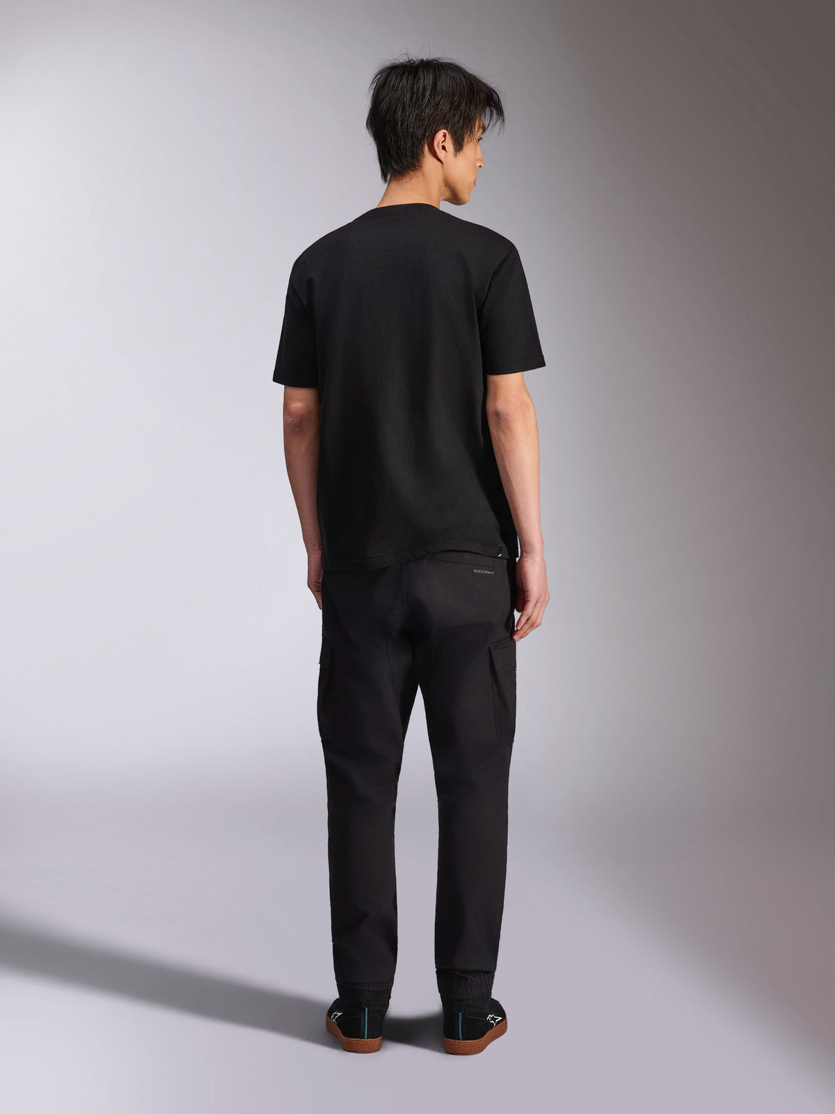 Alpinestars Noka CSF Tee, kurzärmeliges T-Shirt, Schwarz, Rückansicht eines Models, das das T-Shirt mit schwarzer Hose und Sneakern trägt, lässiger Motorsport-Streetwear-Stil