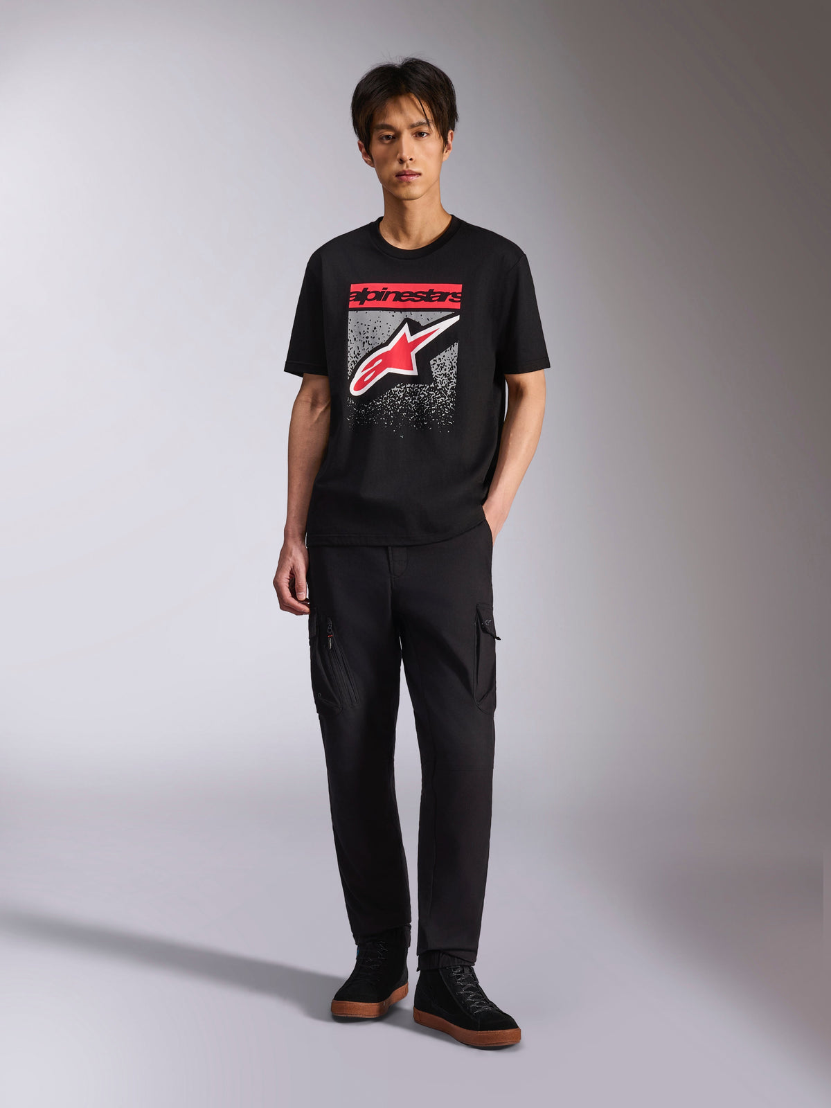 Alpinestars Noka CSF Tee, kurzärmeliges T-Shirt, Schwarz, Astars-Logo auf der Vorderseite mit rot-weißer MX-Verlaufsgrafik, getragen von einem Model mit schwarzer Cargohose, lässige Motorsport-Streetwear